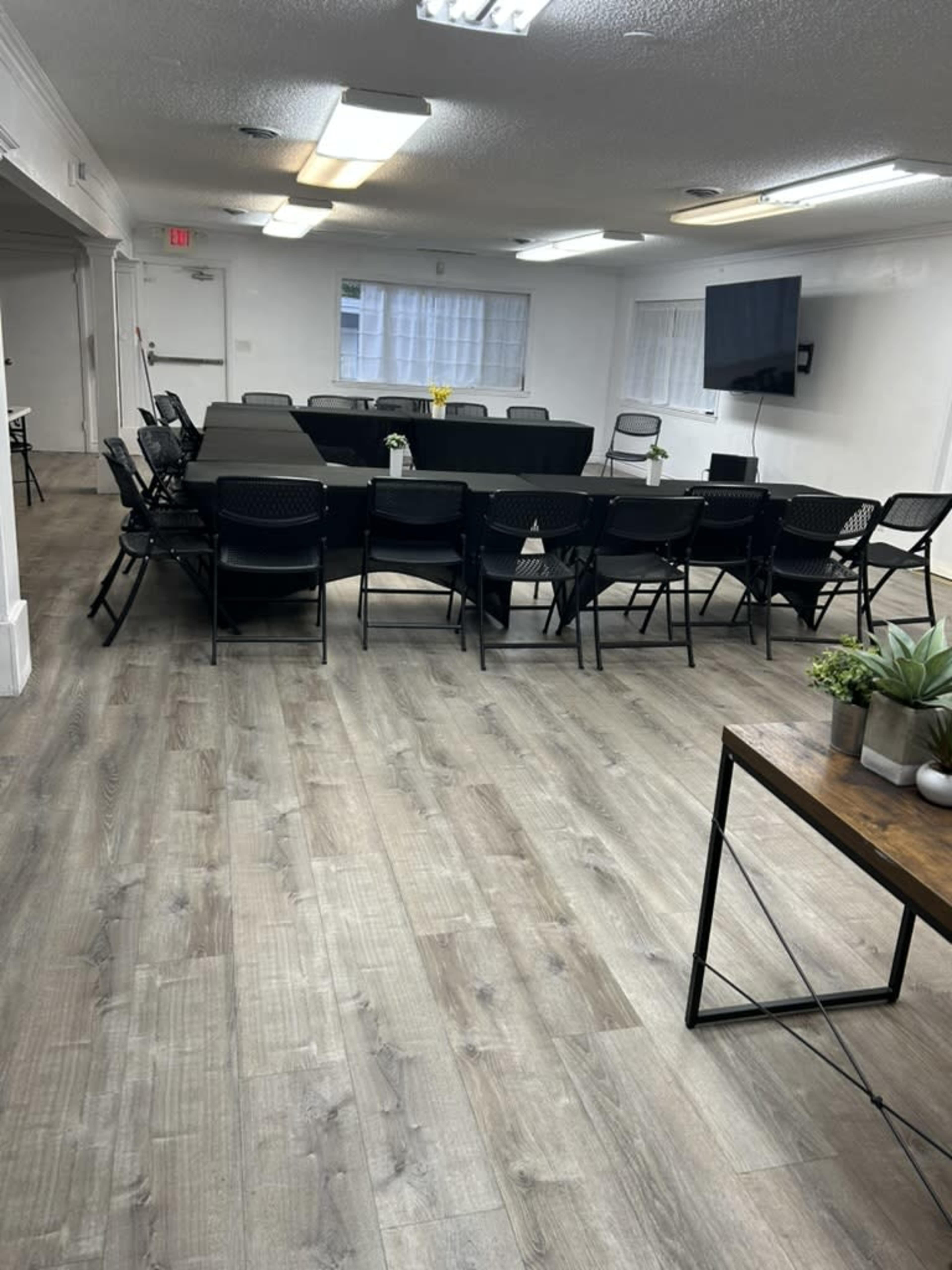 A conference room features a large rectangular table surrounded by chairs, with a television on the wall and a window allowing natural light.