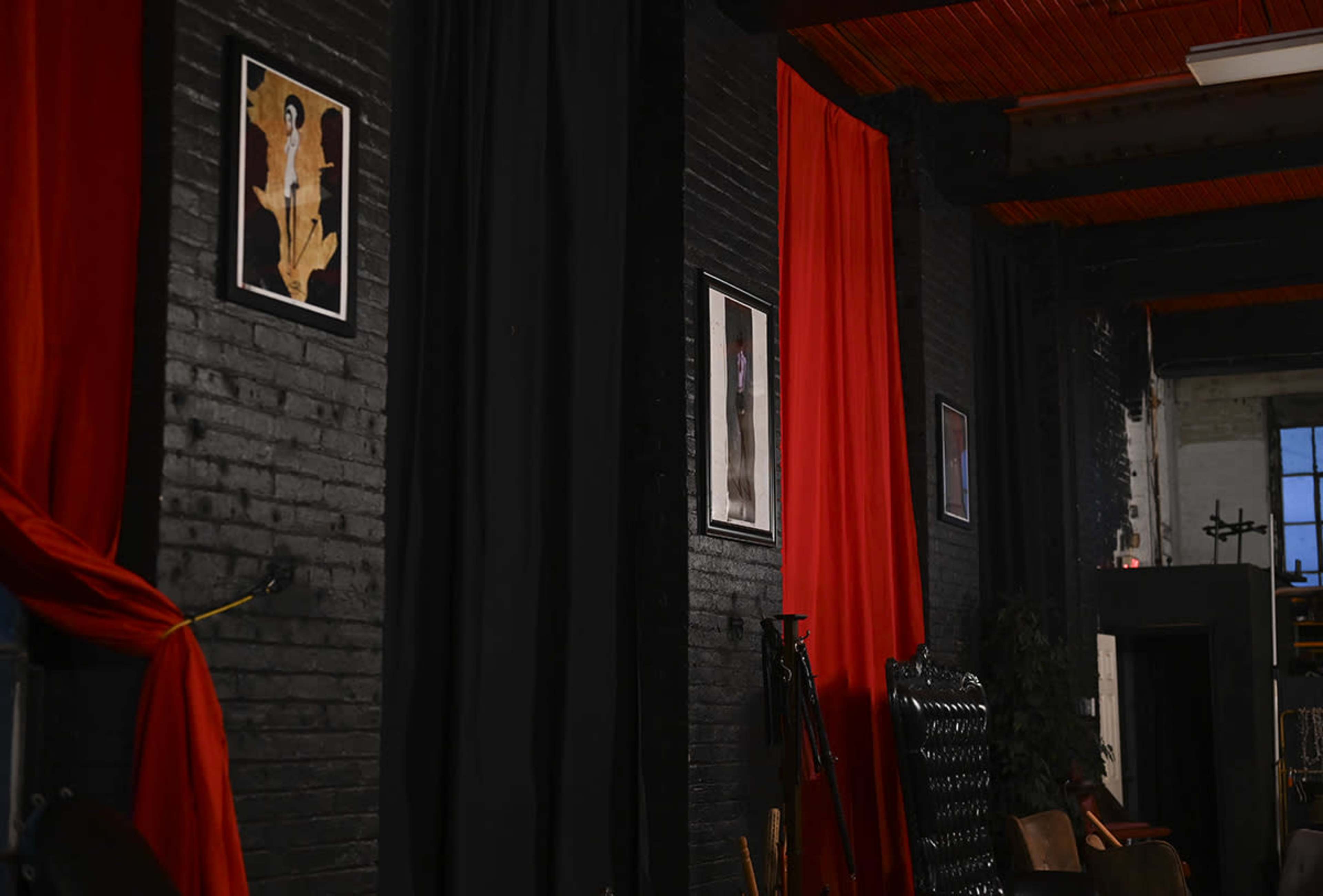 The image shows a dimly lit interior wall featuring black and red drapes along with framed artworks arranged on the brick surface.