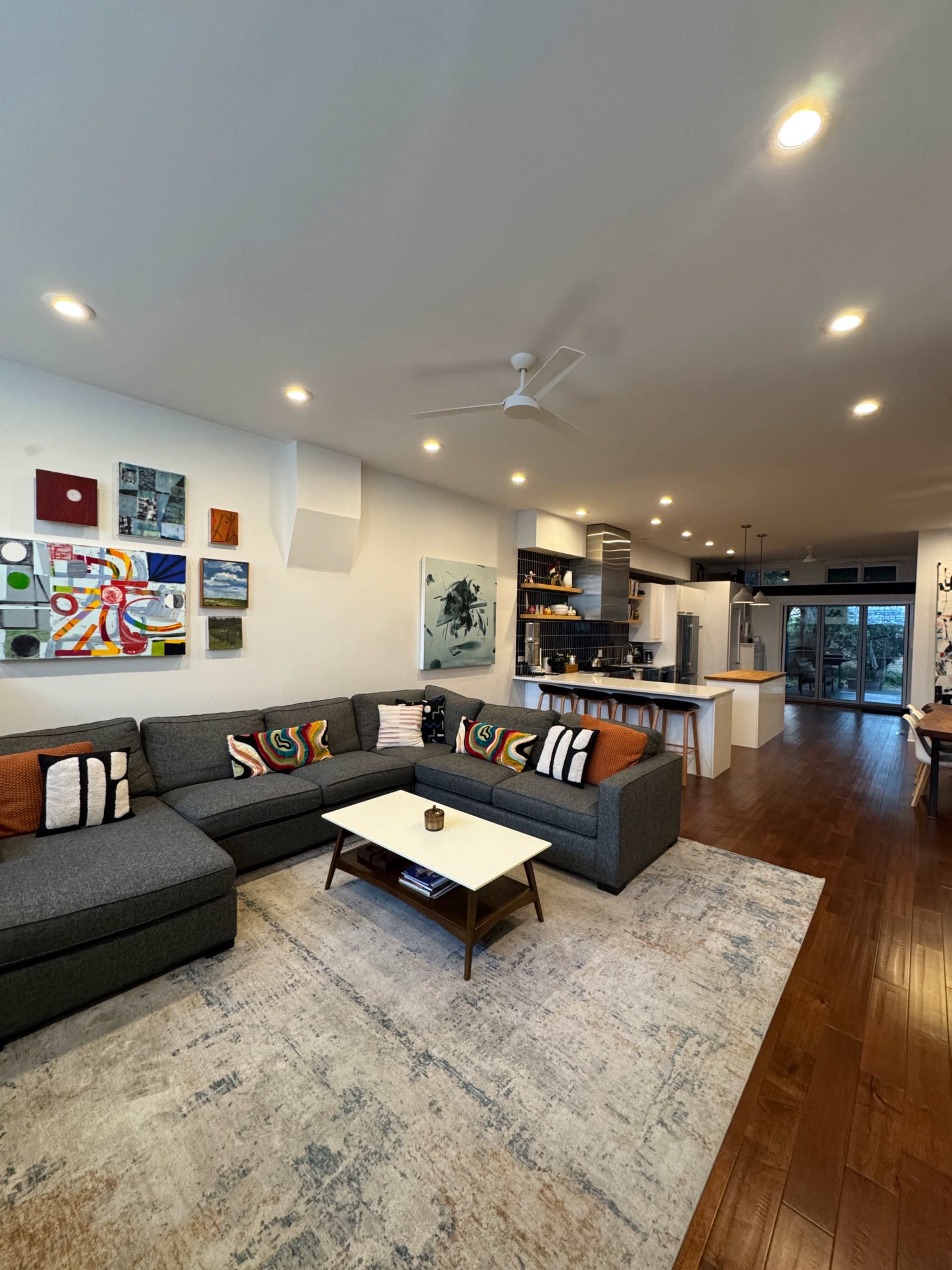 The image shows a modern living room featuring a large gray sectional sofa, a central coffee table, a patterned area rug, and an open kitchen in the background with a sliding glass door leading to an outdoor space.