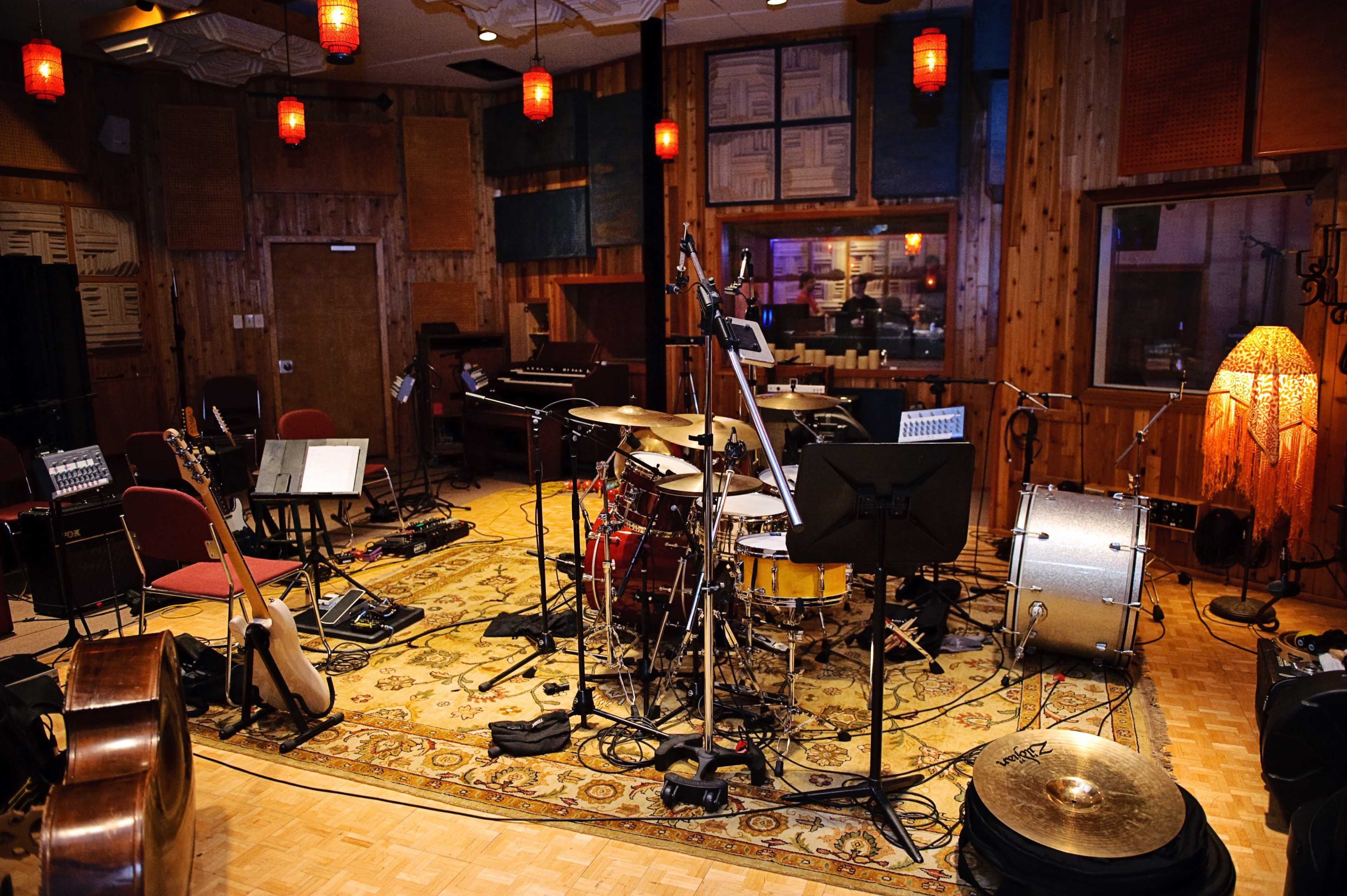 A music studio is set up with a drum kit, guitars, and various microphones, surrounded by wooden walls and ambient lighting.