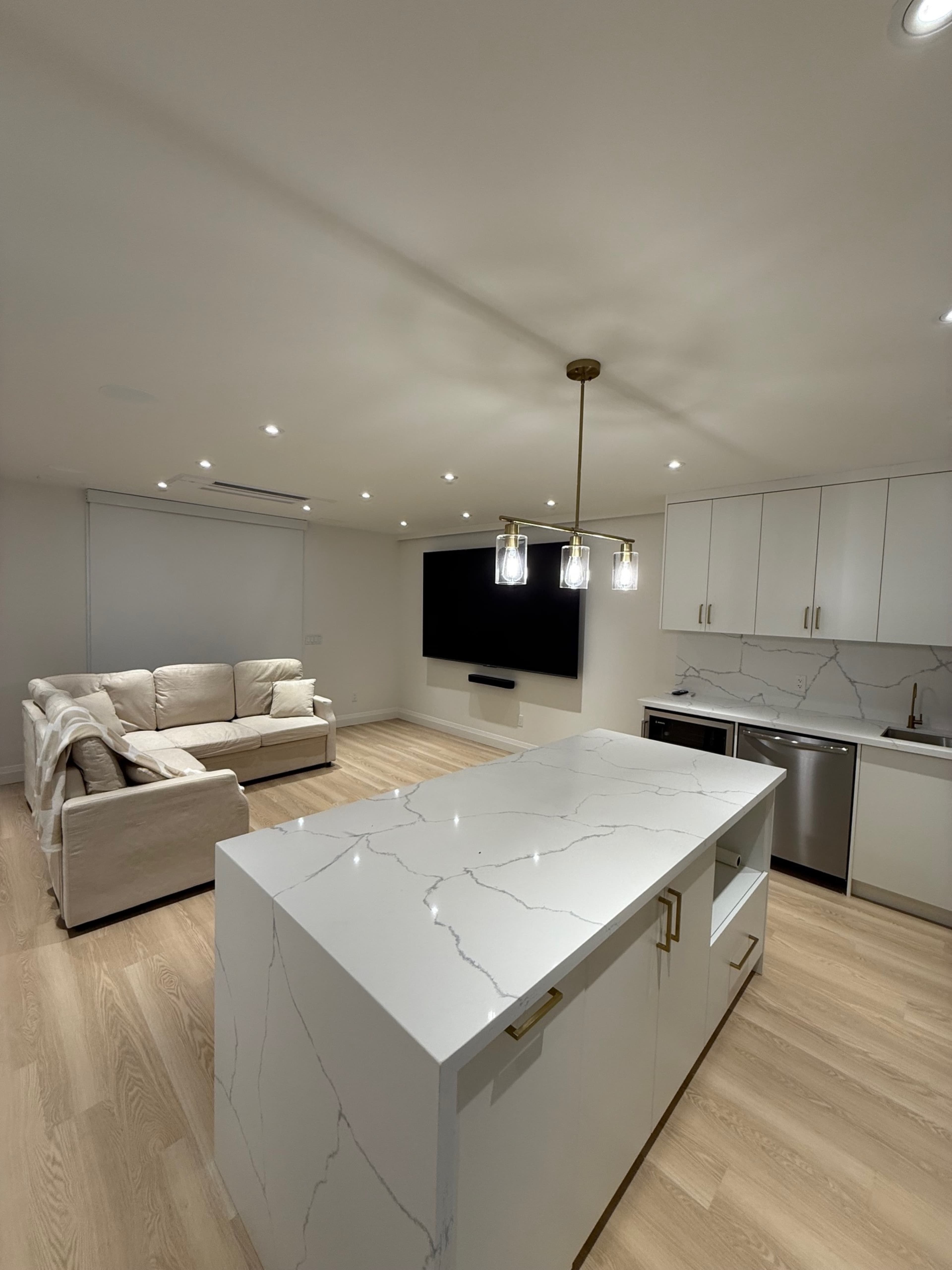 The image shows a modern kitchen and living area featuring a large island with a white marble countertop, a beige couch, and a wall-mounted television.