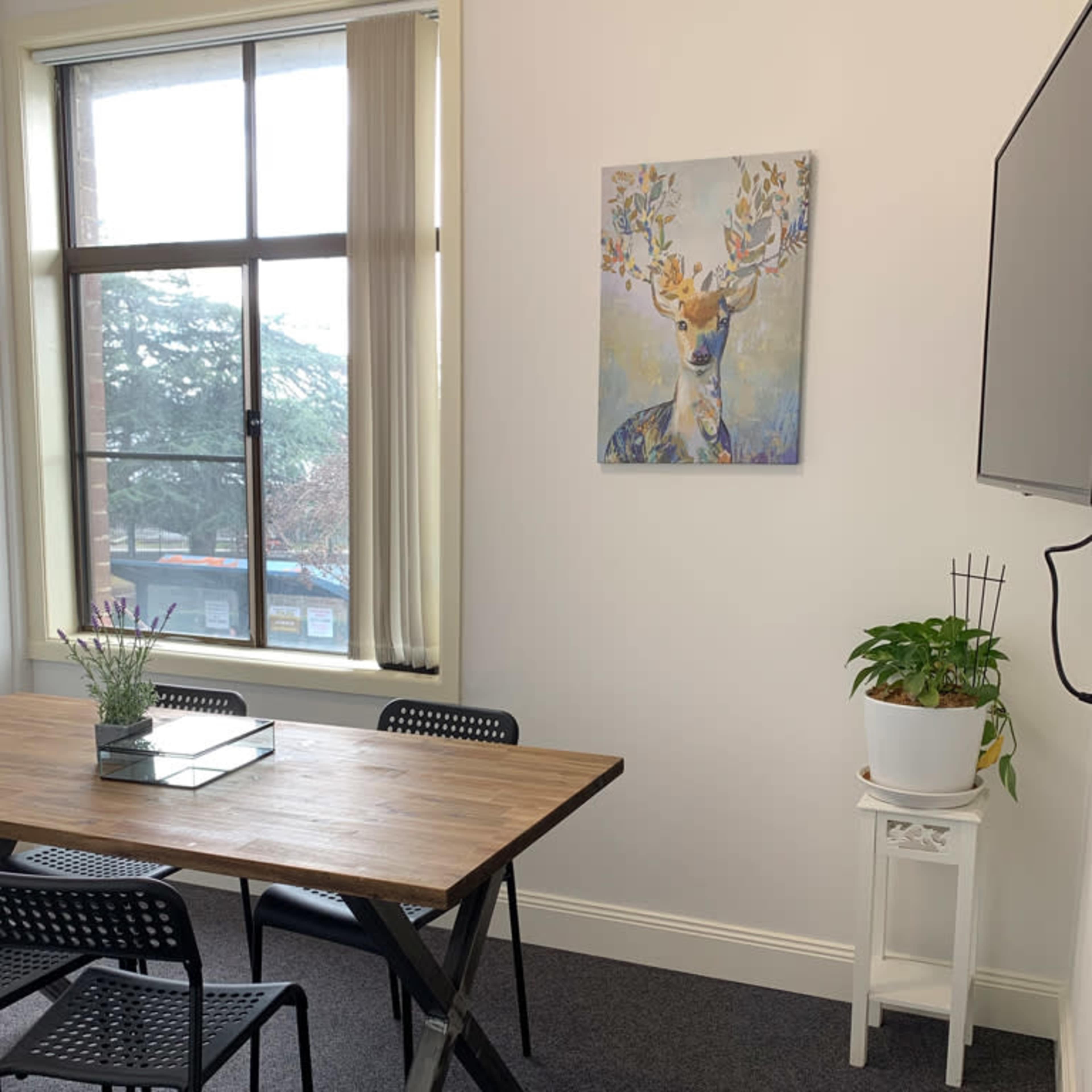 A small meeting room with a wooden table, black chairs, a deer painting on the wall, and a plant on a stand near a window.