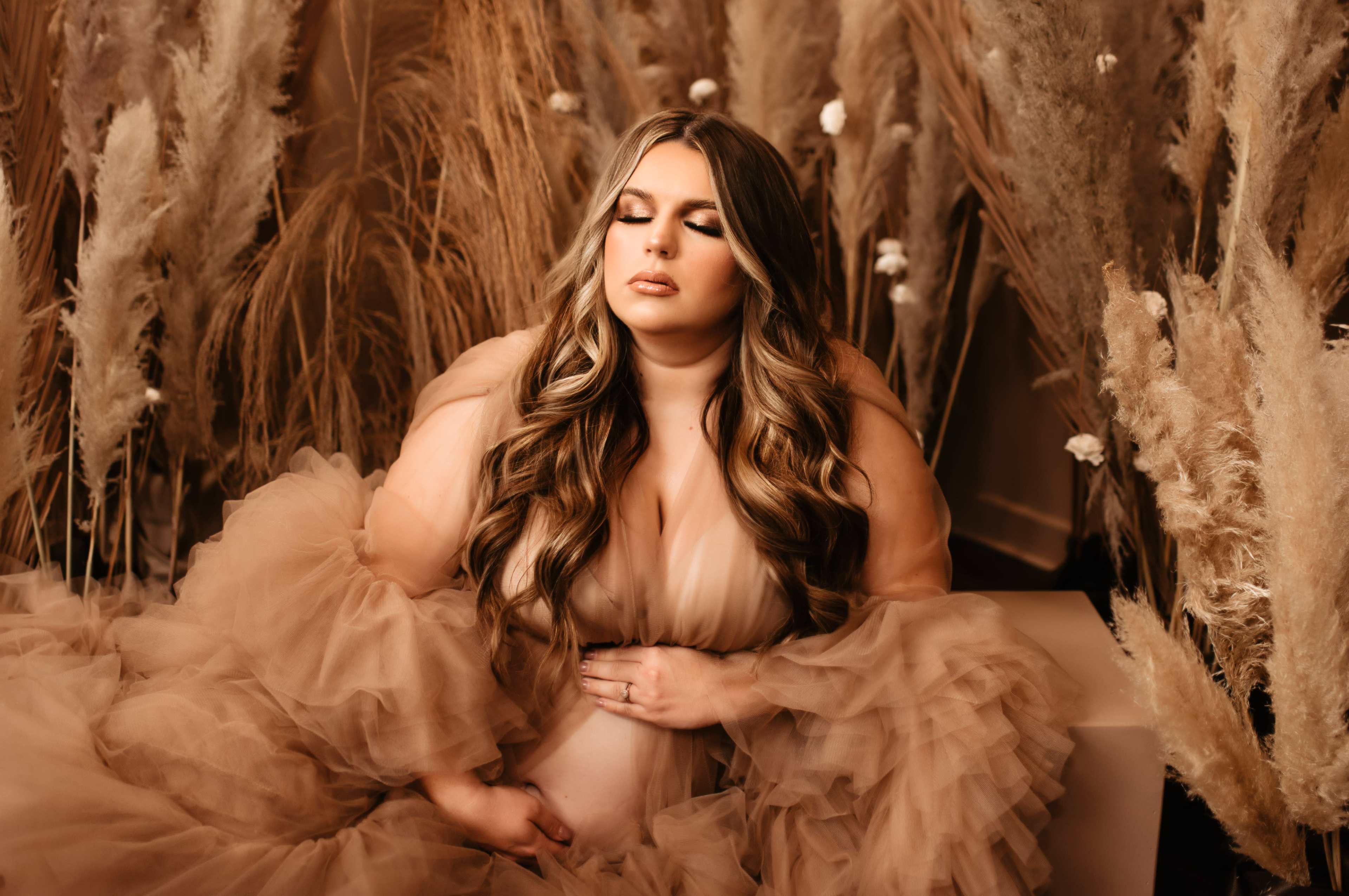 A pregnant woman in a flowing tan gown poses serenely among tall dried grasses and soft floral accents.