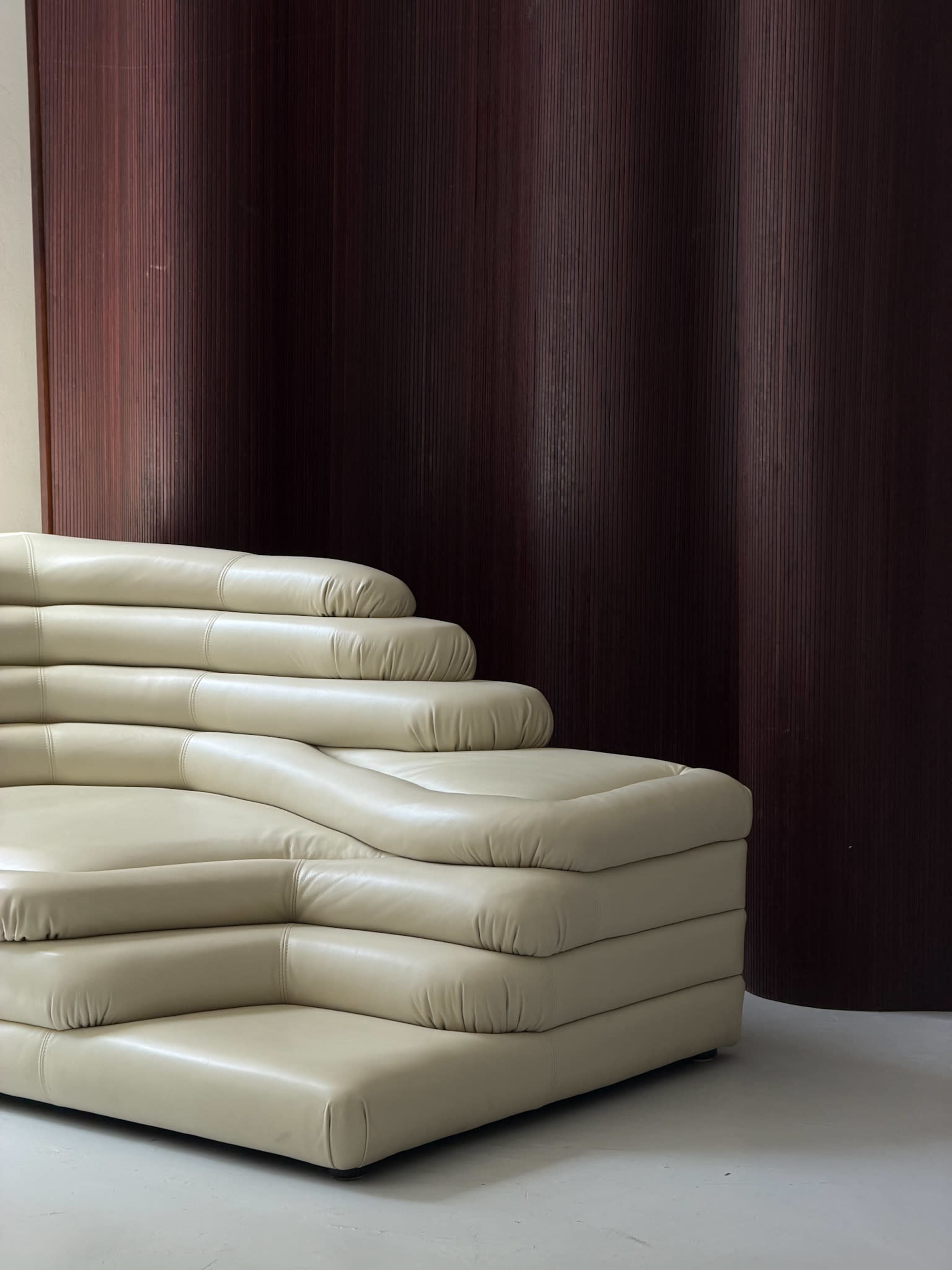 The image shows a modern, cream-colored sectional sofa with a wave-like design positioned against a textured, dark red wall.