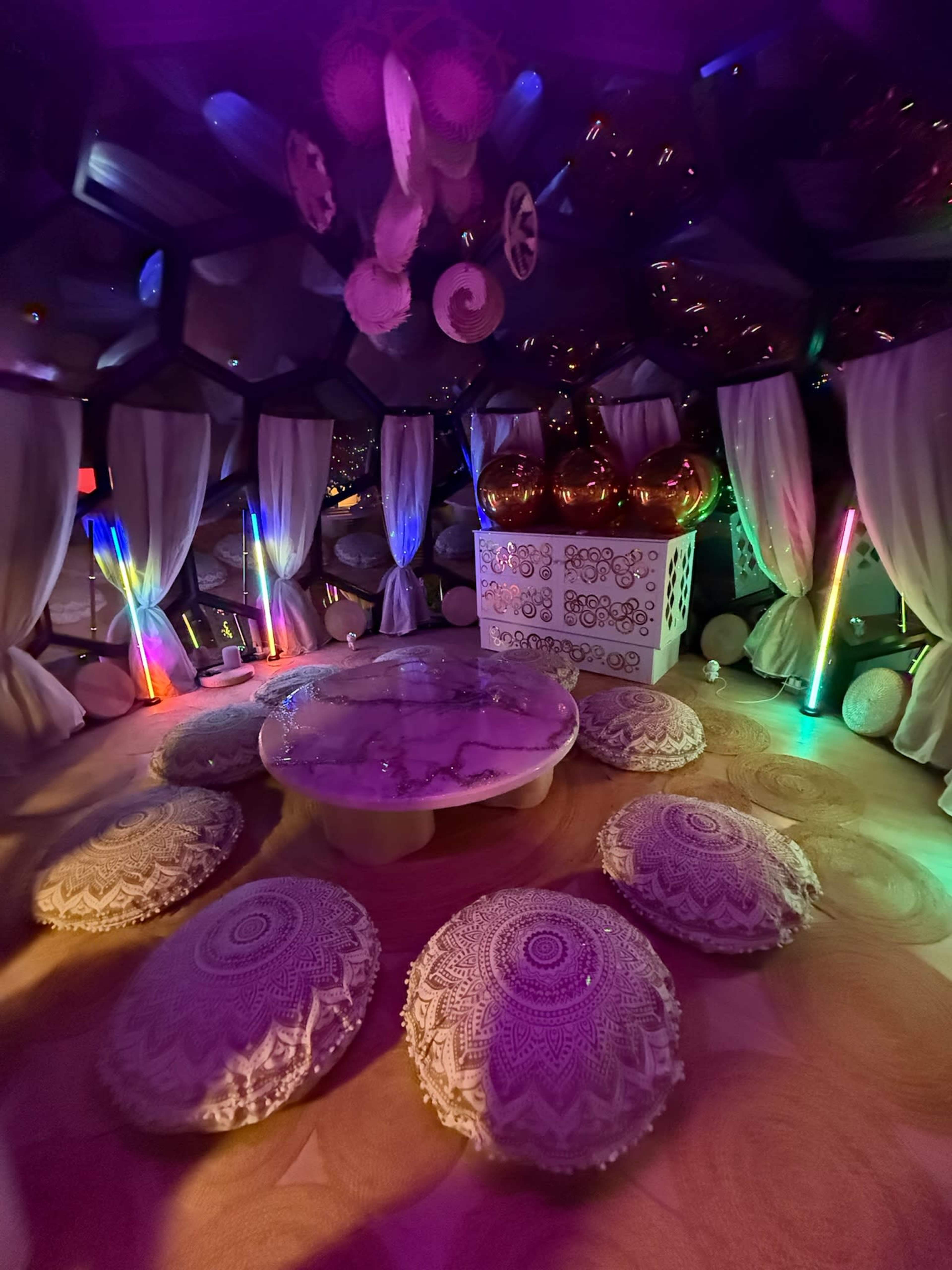 The image shows a dimly lit lounge with a round table in the center, surrounded by upholstered floor cushions and colorful LED lights enhancing the atmosphere.
