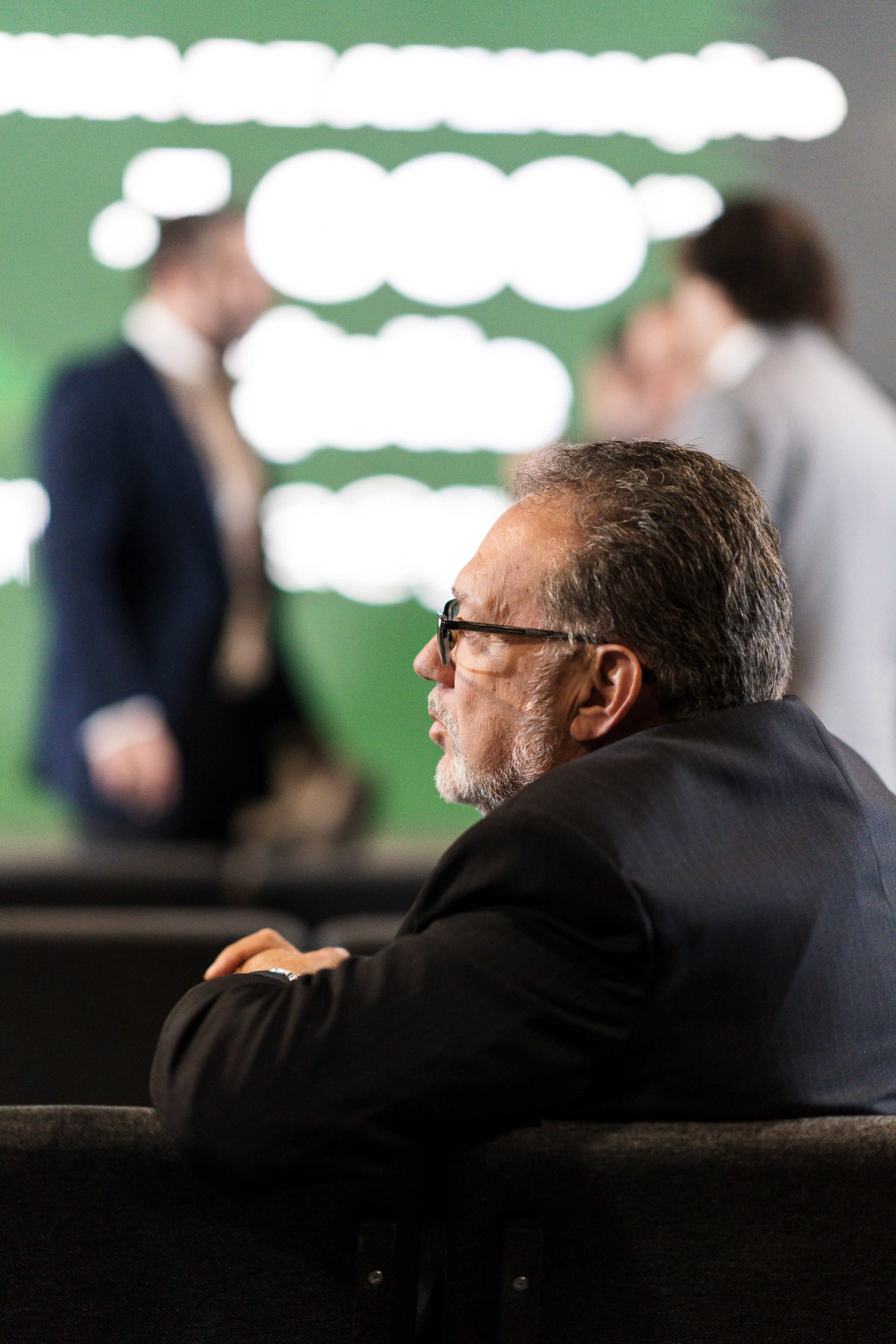 A man in a suit sits on a couch with blurred figures engaging in conversation in the background.