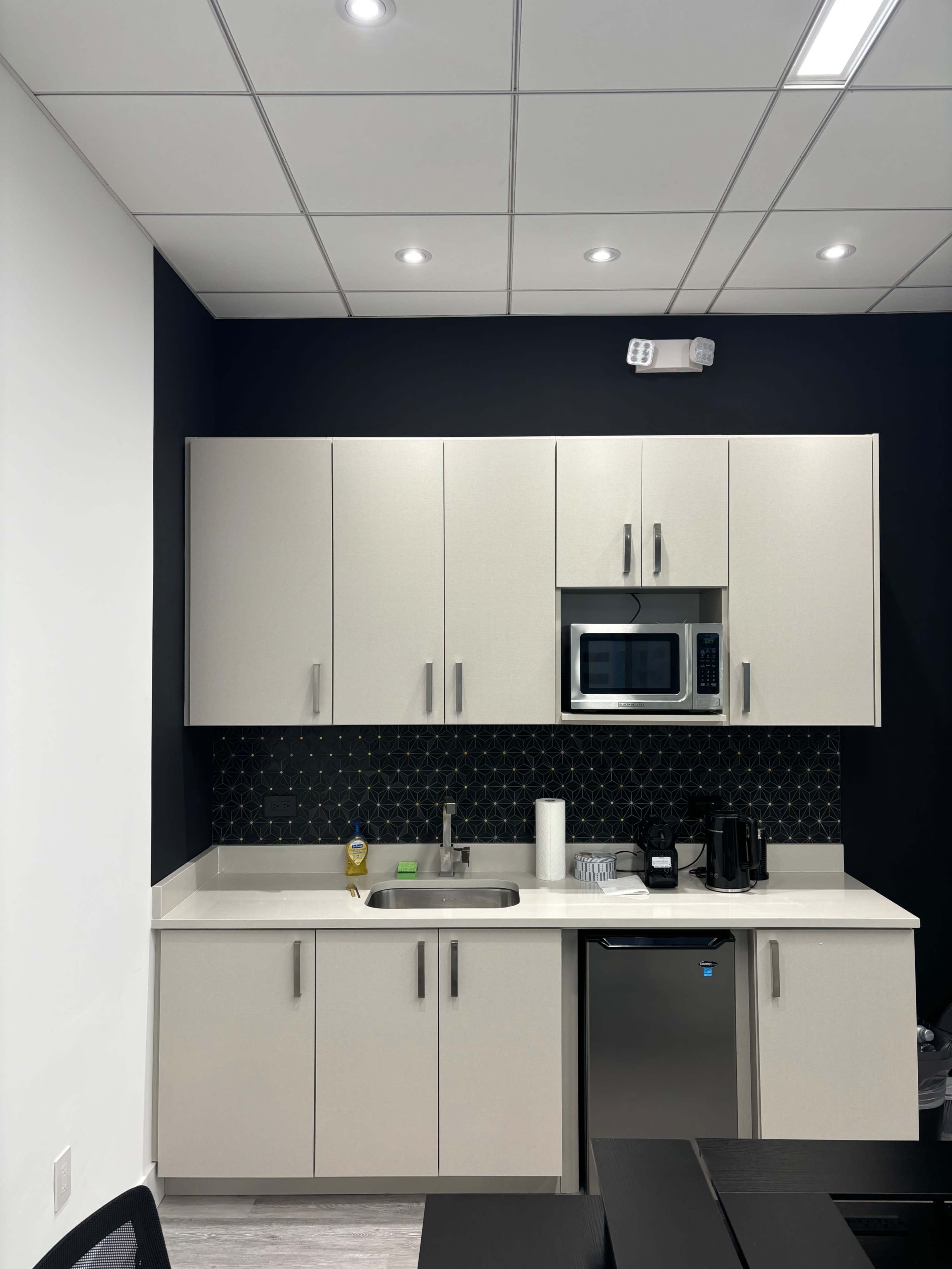 A modern kitchen with light-colored cabinets, a stainless steel microwave, a sink, and a small refrigerator, set against a dark accent wall.