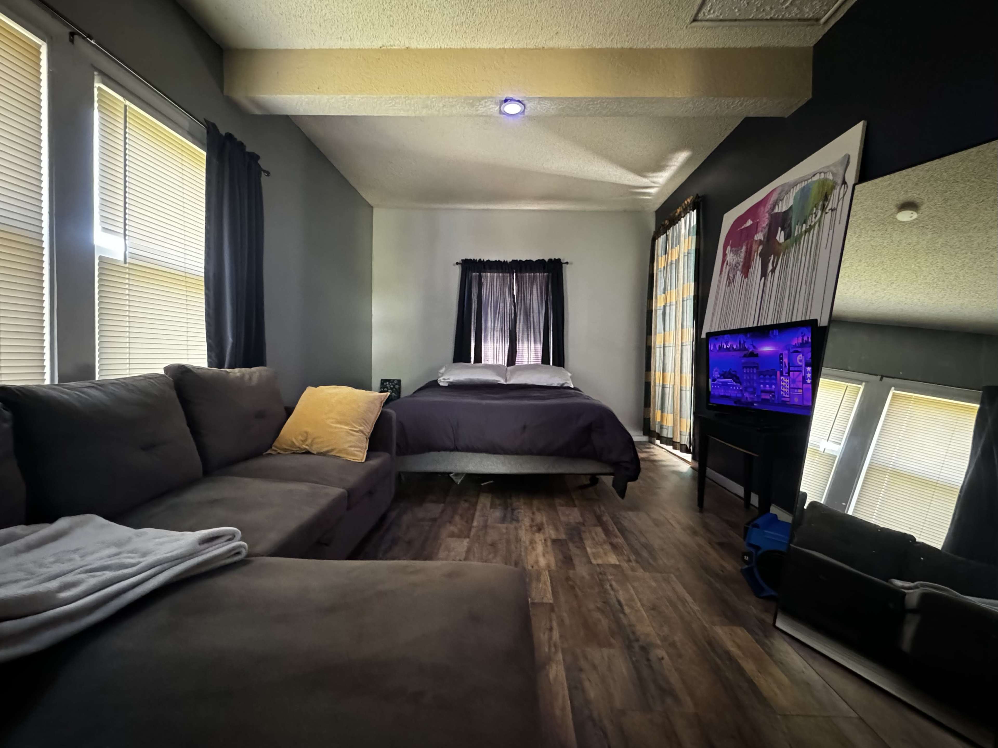 The image shows a compact bedroom with a bed, a sofa, a television on a stand, and large windows with curtains.