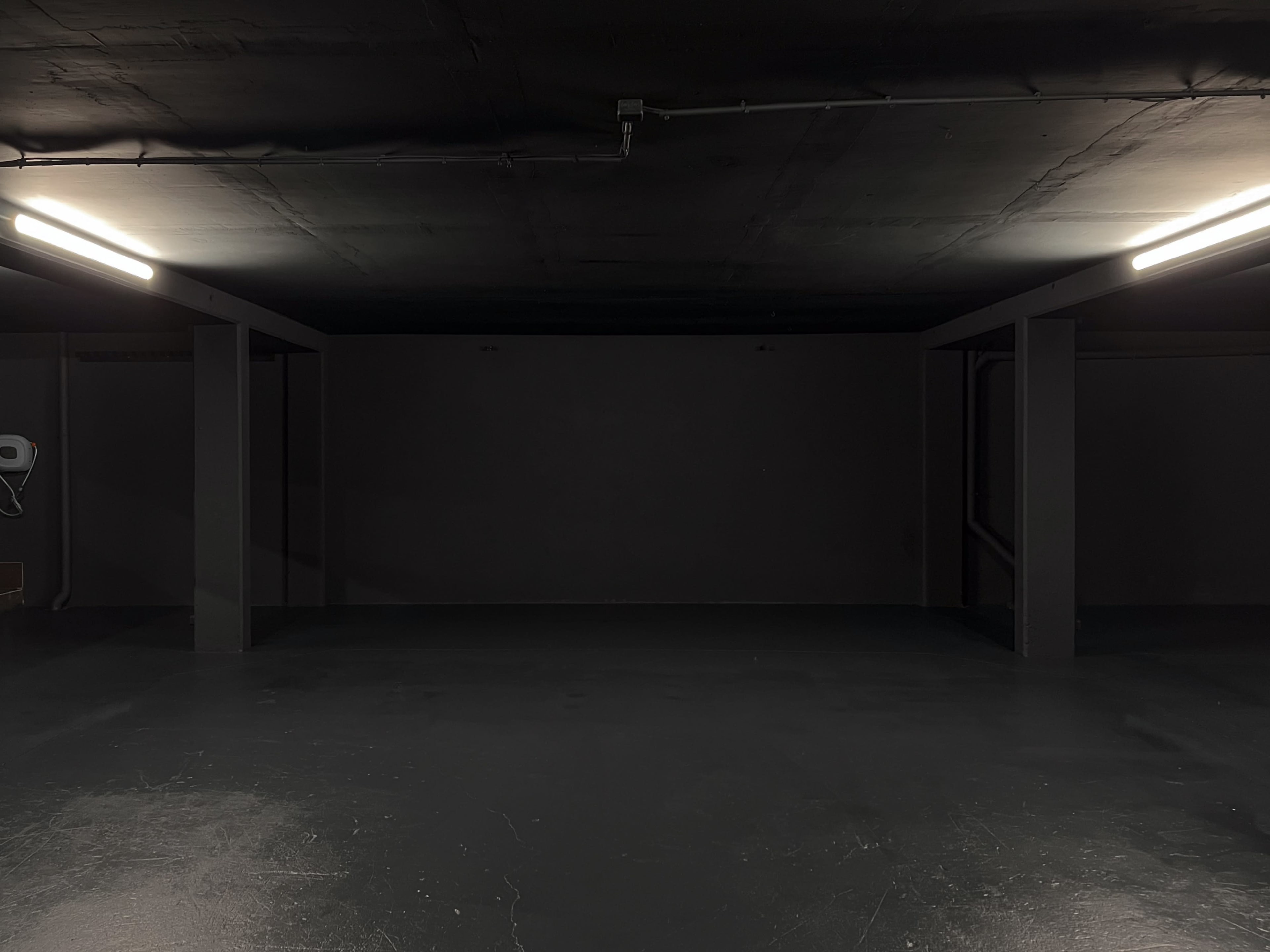 The image shows an empty, dimly lit underground room with a concrete floor and dark walls, featuring two fluorescent light fixtures.