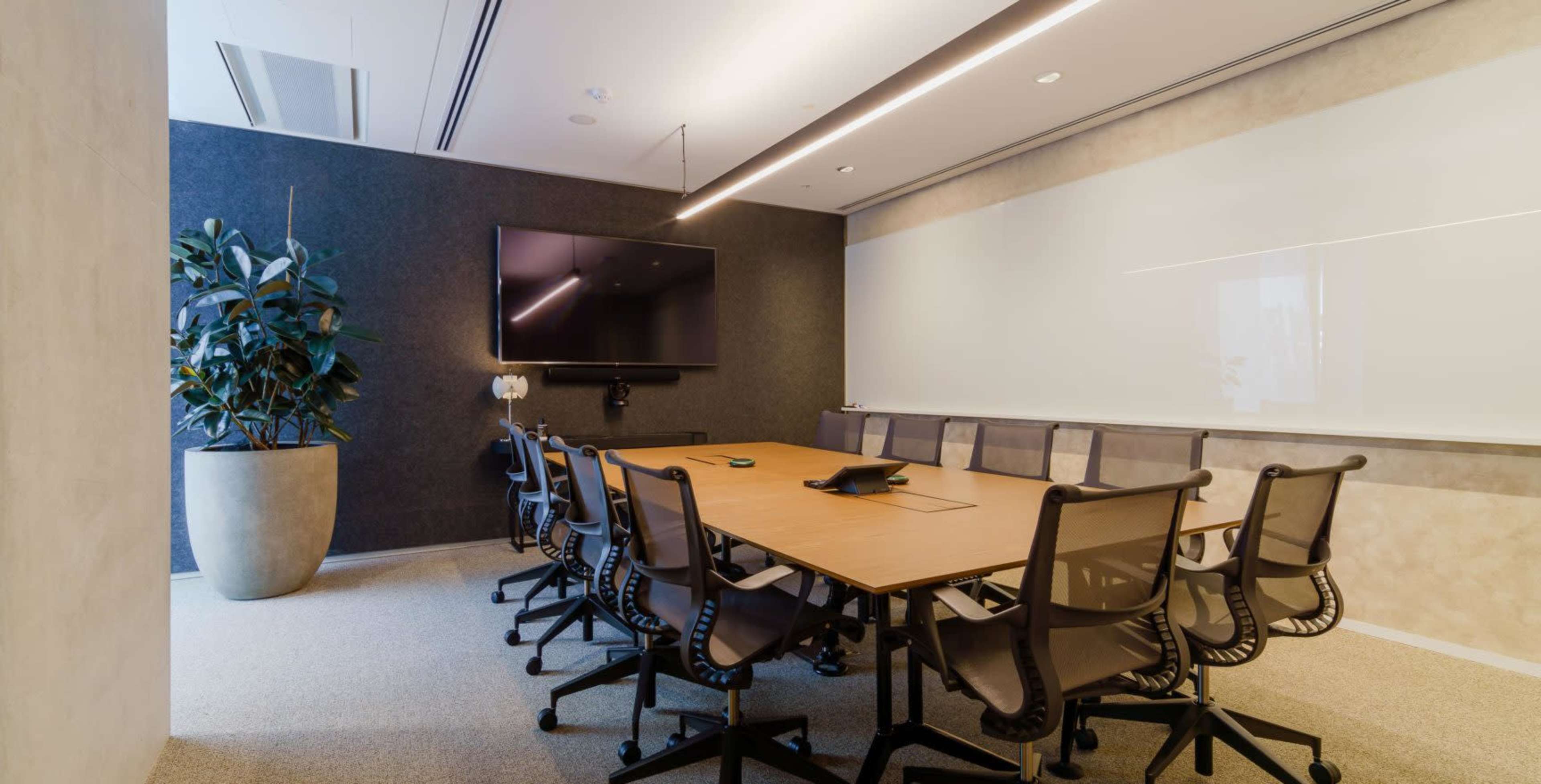 A modern conference room features a large table surrounded by ergonomic chairs, a television screen mounted on the wall, and a potted plant in the corner.
