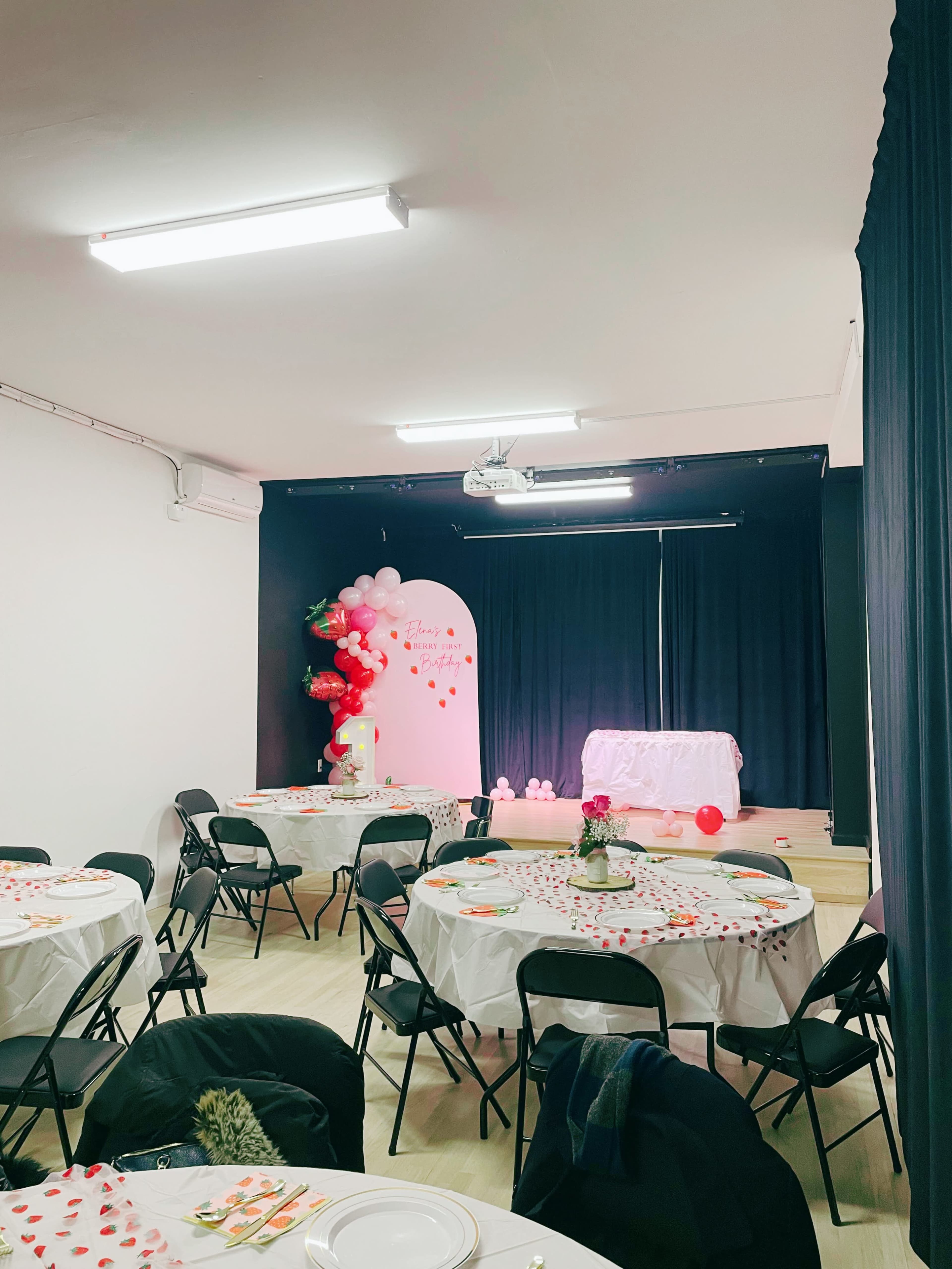 A decorated event space features round tables with white tablecloths, each set for dining, and a stage area adorned with balloons and a white tablecloth backdrop.