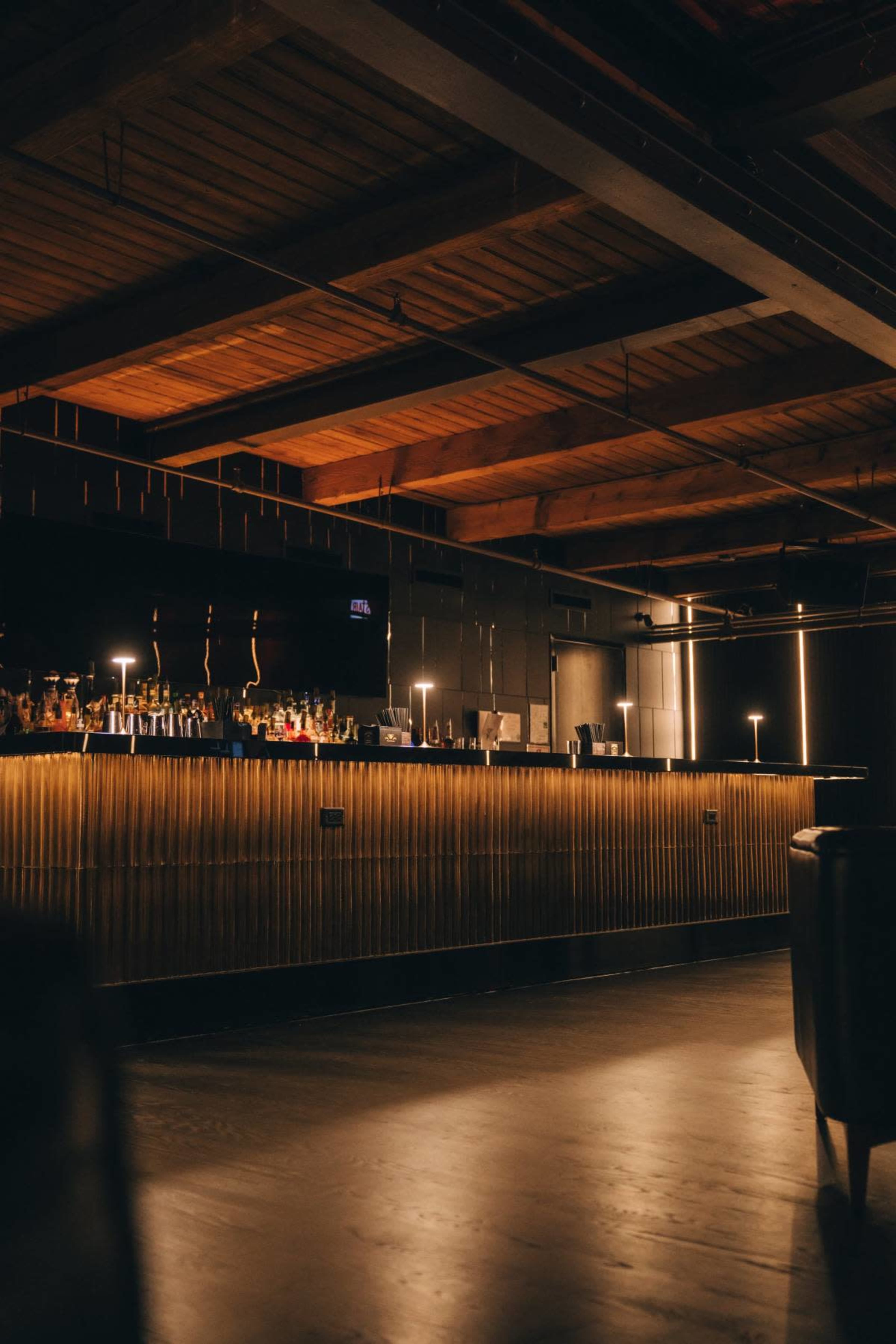 The image shows a dimly lit bar area with a sleek wooden counter and shelves filled with various bottles in the background.