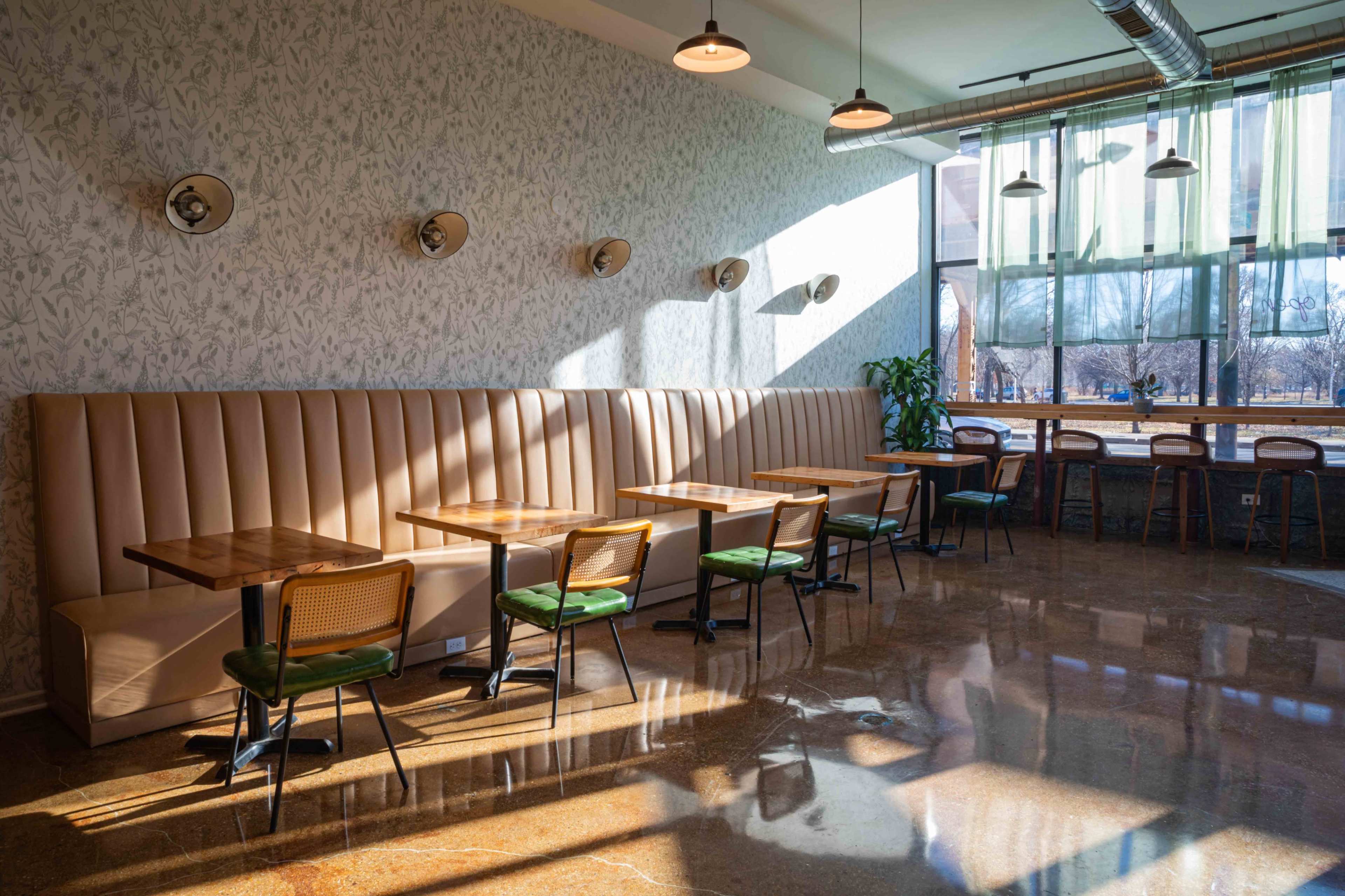 The image shows a spacious café interior with a combination of green-upholstered tables and chairs, a long padded bench along one wall, and large windows letting in natural light.