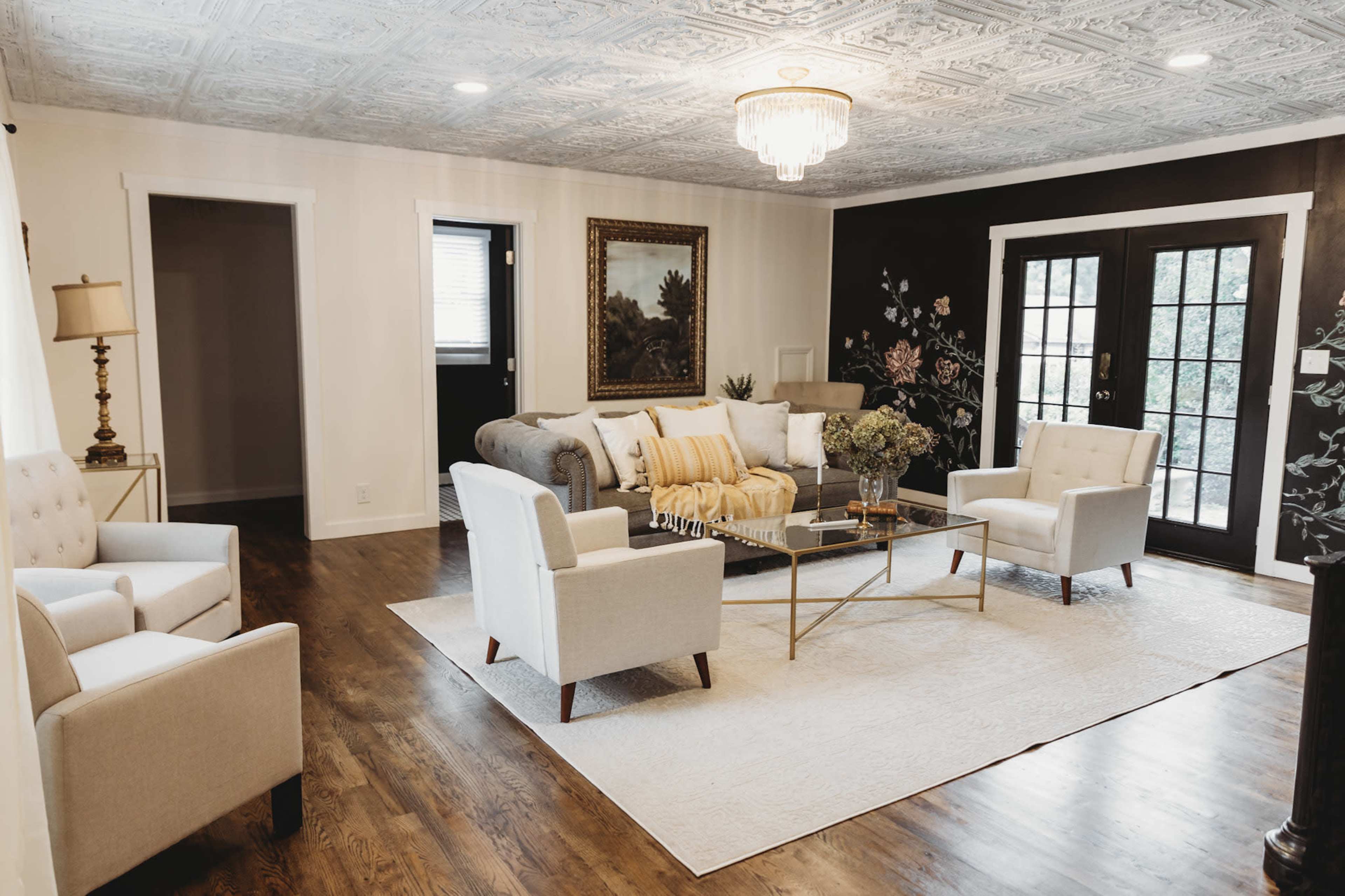 A modern living room with a gray sofa, two white armchairs, a glass coffee table, and large windows leading to an outdoor area.