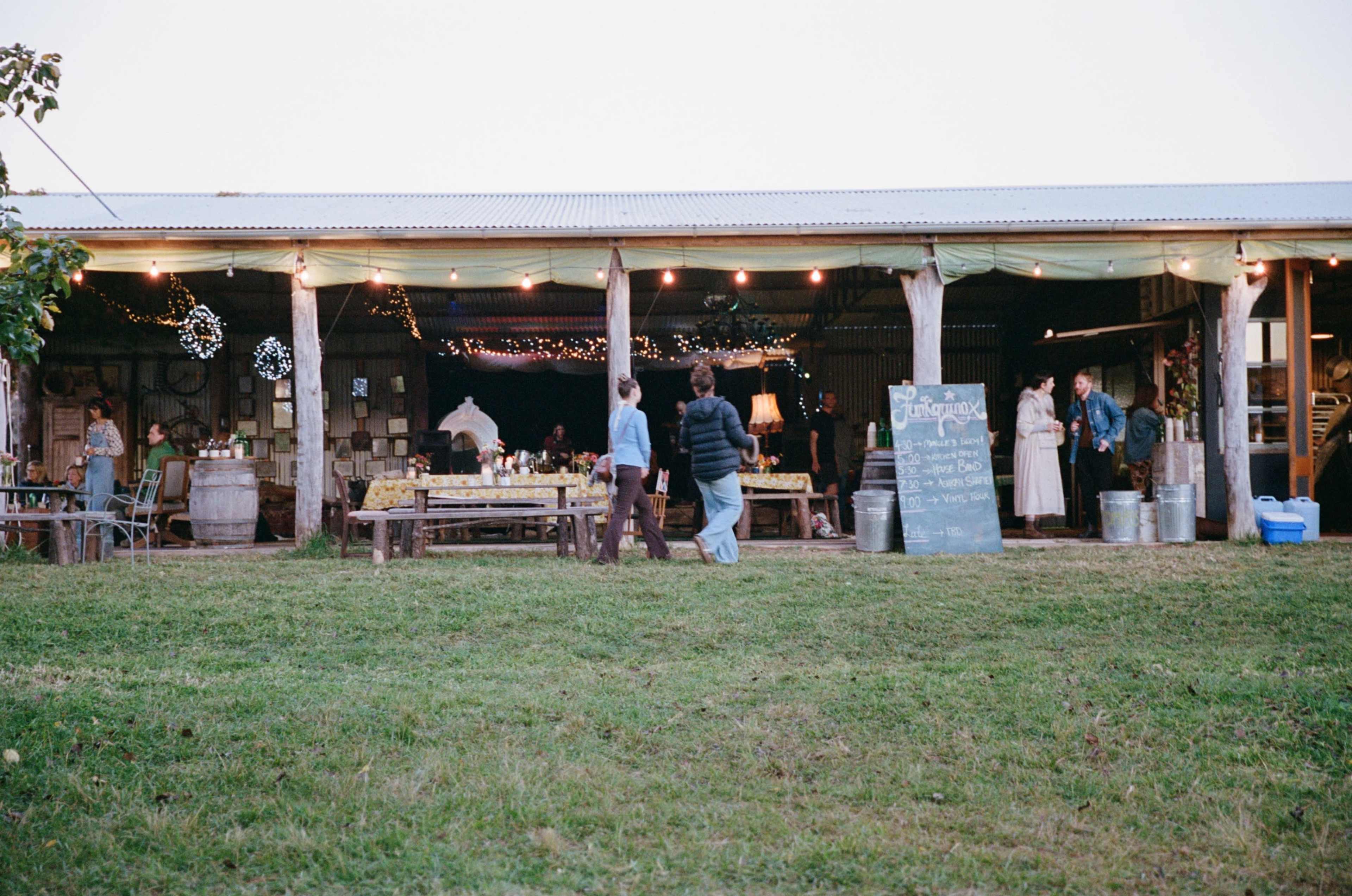 A group of people strolls through a barn decorated with lights, tables, and a chalkboard sign amidst an outdoor gathering.