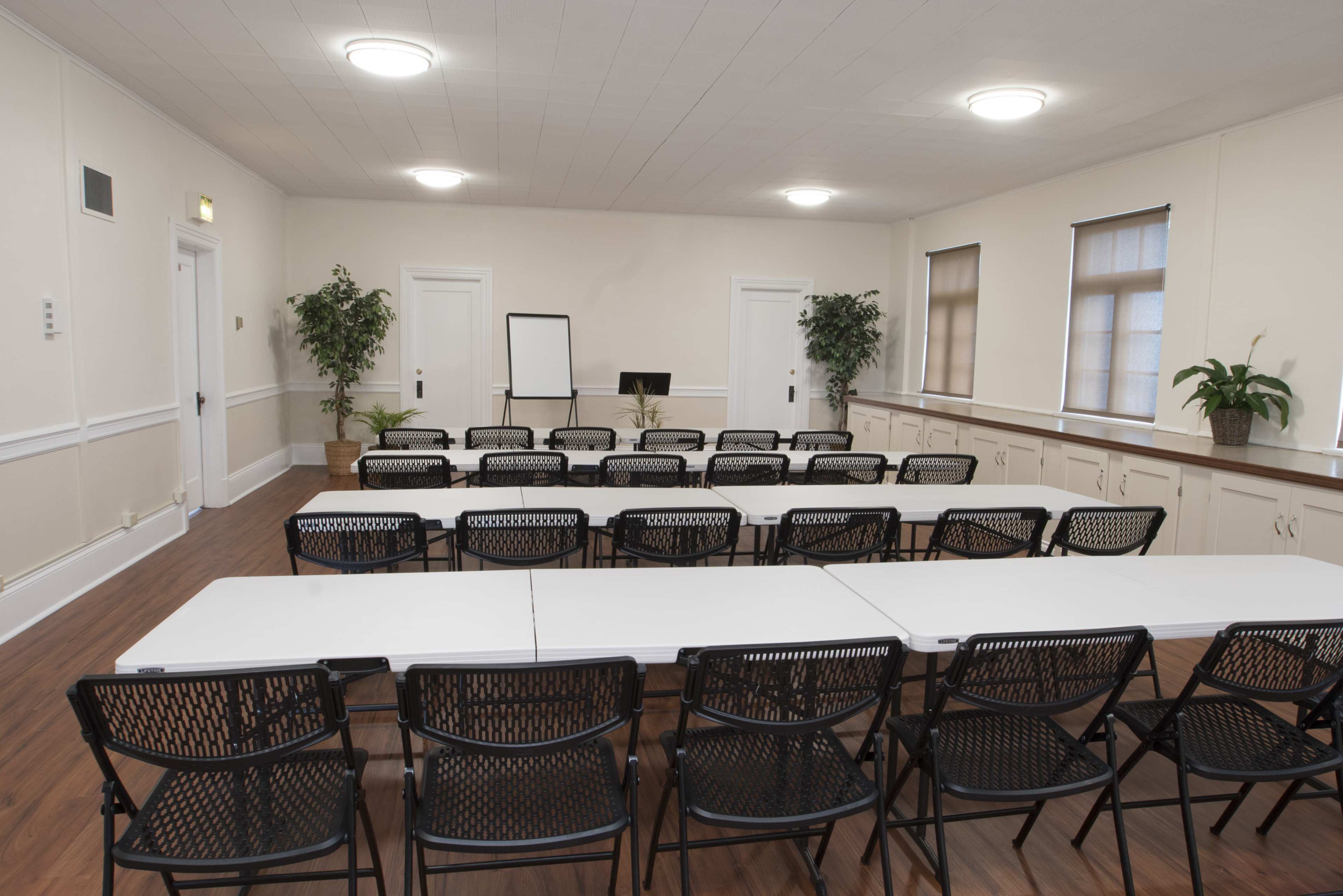 A conference room is arranged with rows of black chairs facing a central table, flanked by potted plants and a whiteboard.