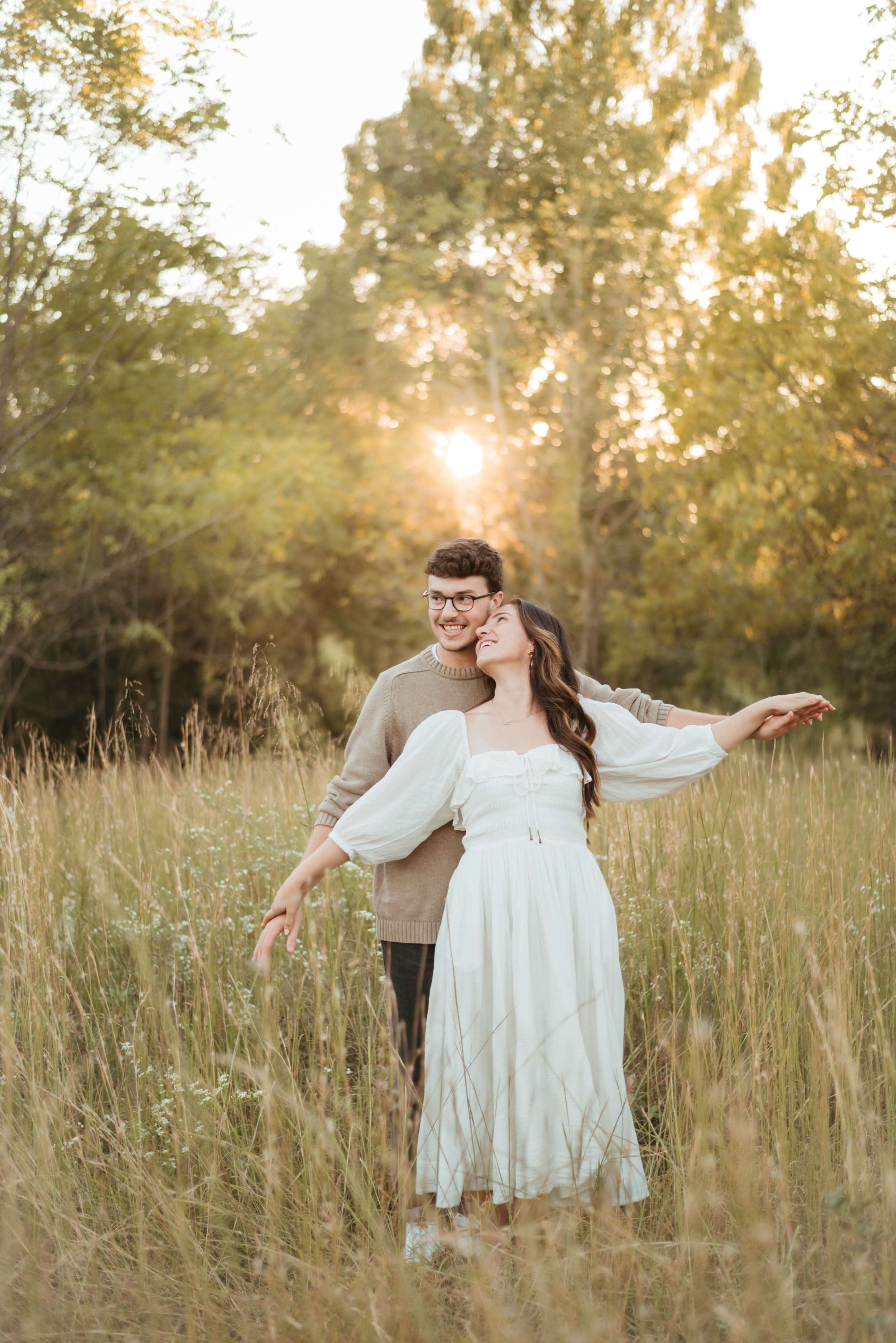 A couple stands in a field of tall grass, with the sun setting behind them, as they share a joyful moment together.