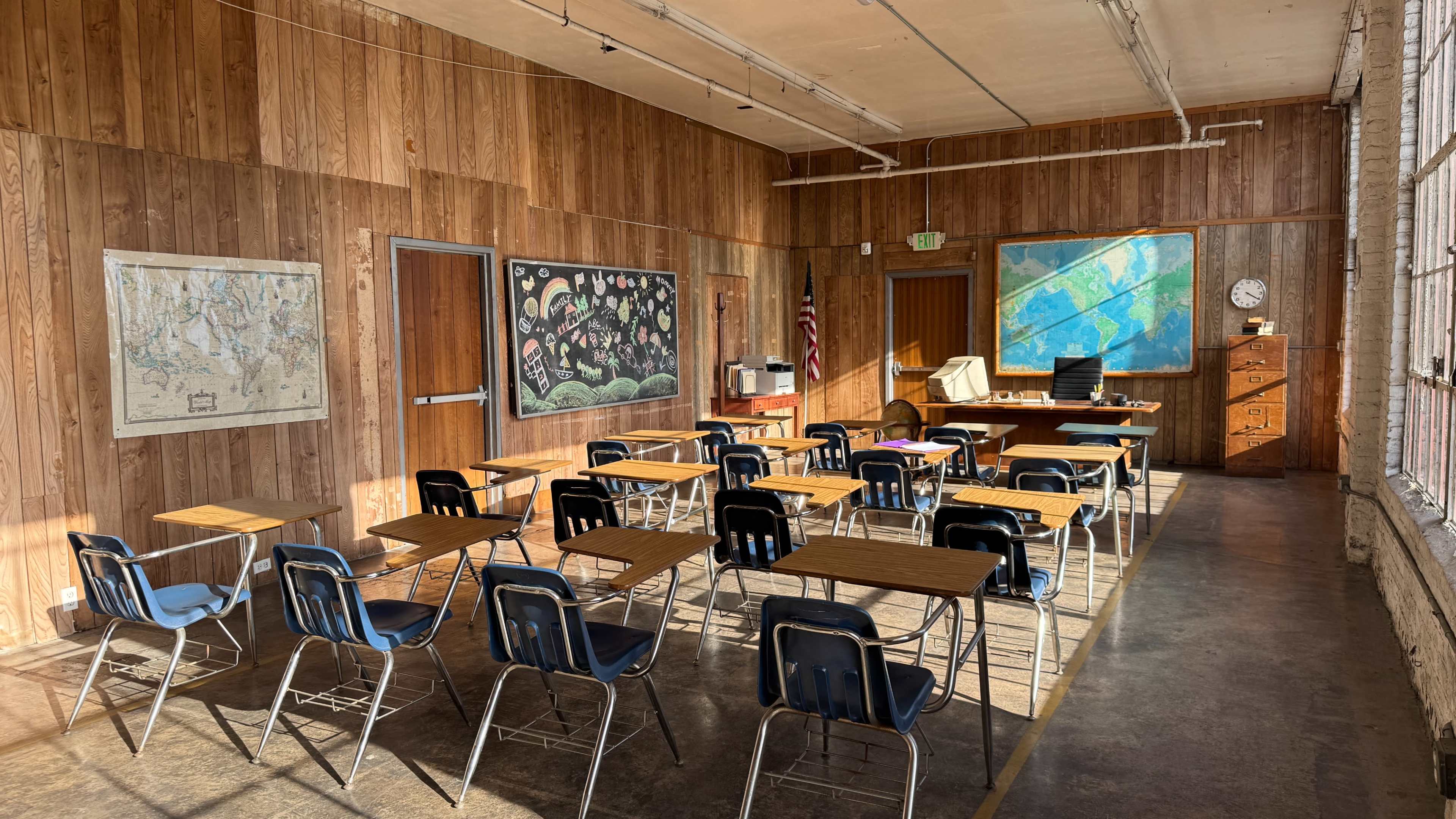 The image depicts a school classroom with wooden walls, multiple desks arranged in rows, and a teacher's desk at the front near maps and a clock.