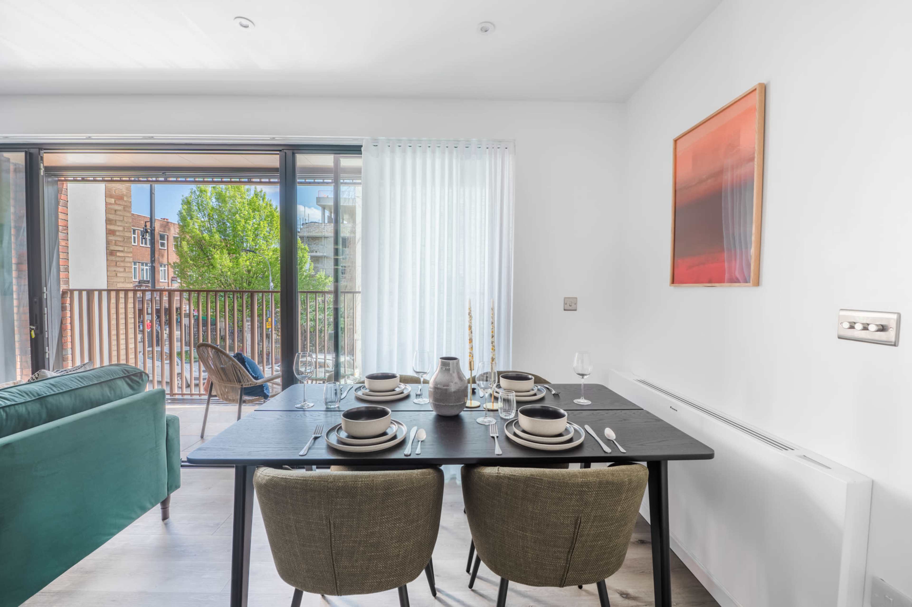 A dining table is set with dishes and glassware, situated in a brightly lit room that opens to a balcony with greenery visible outside.