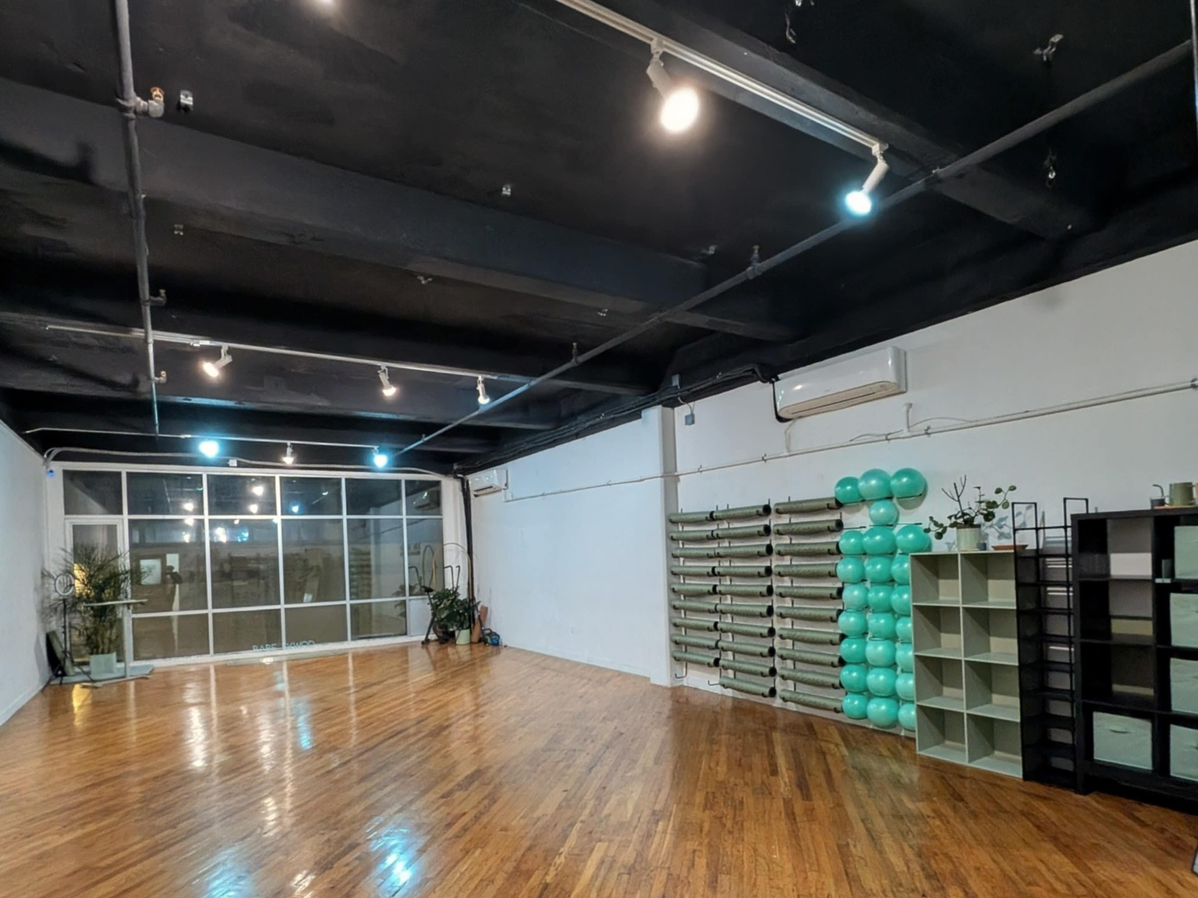 The image shows a spacious, empty fitness studio with wooden flooring, large windows, and equipment neatly arranged along one wall.