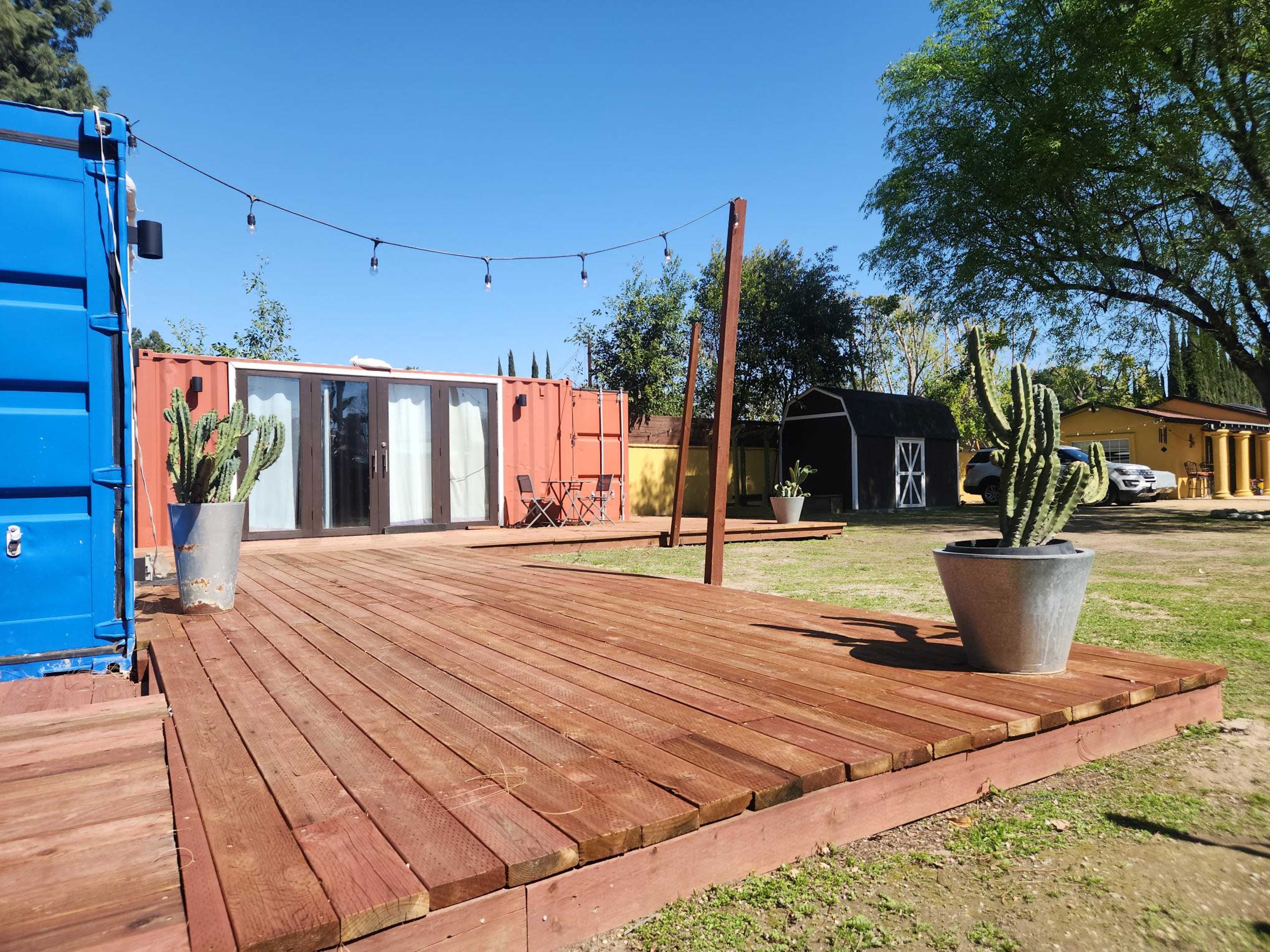 The image shows a wooden deck with potted cacti in the foreground and a shipping container structure in the background, alongside a parked vehicle and string lights overhead.