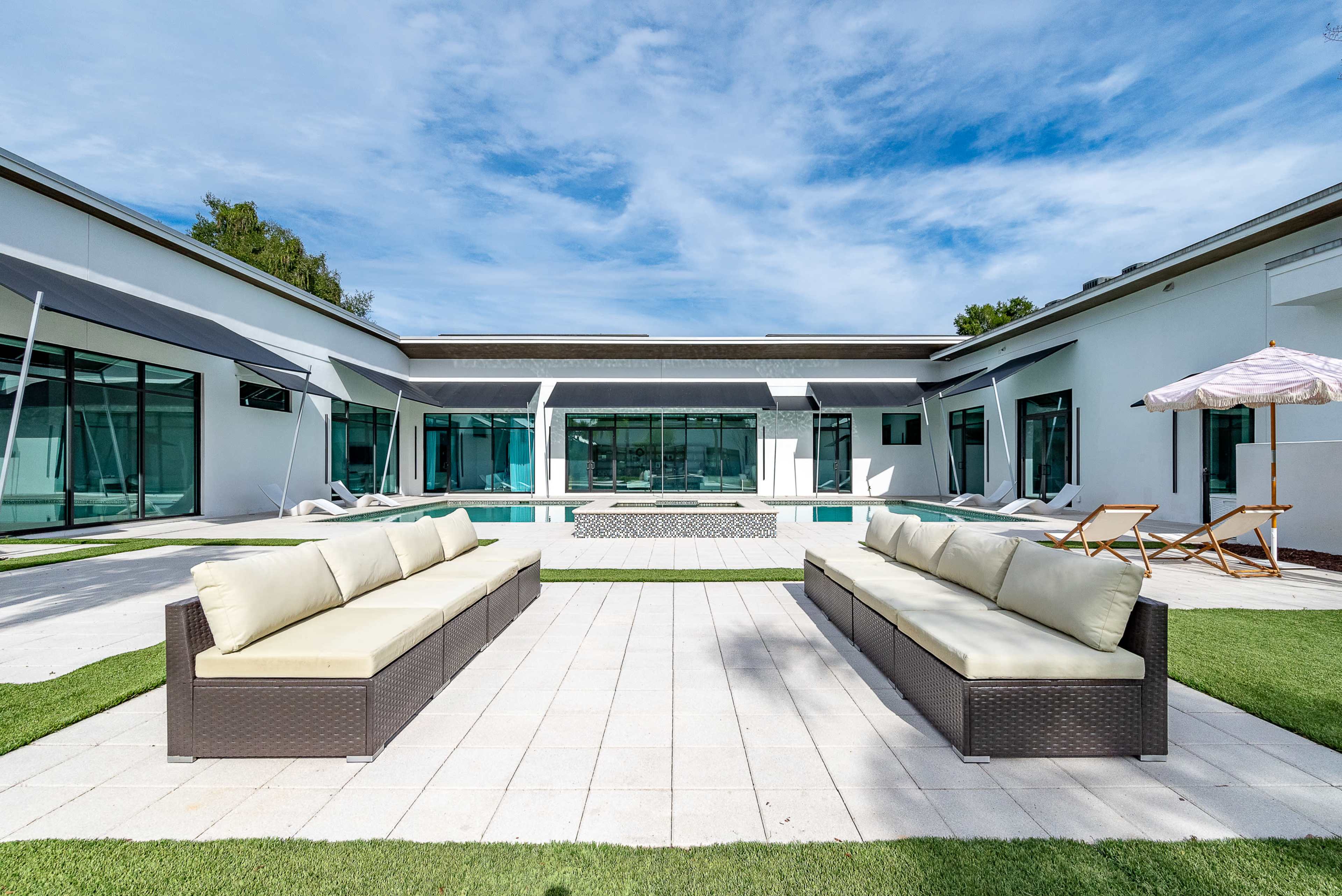 The image shows a modern outdoor area with two cream-colored sofas facing a rectangular swimming pool surrounded by a spacious patio and glass walls.