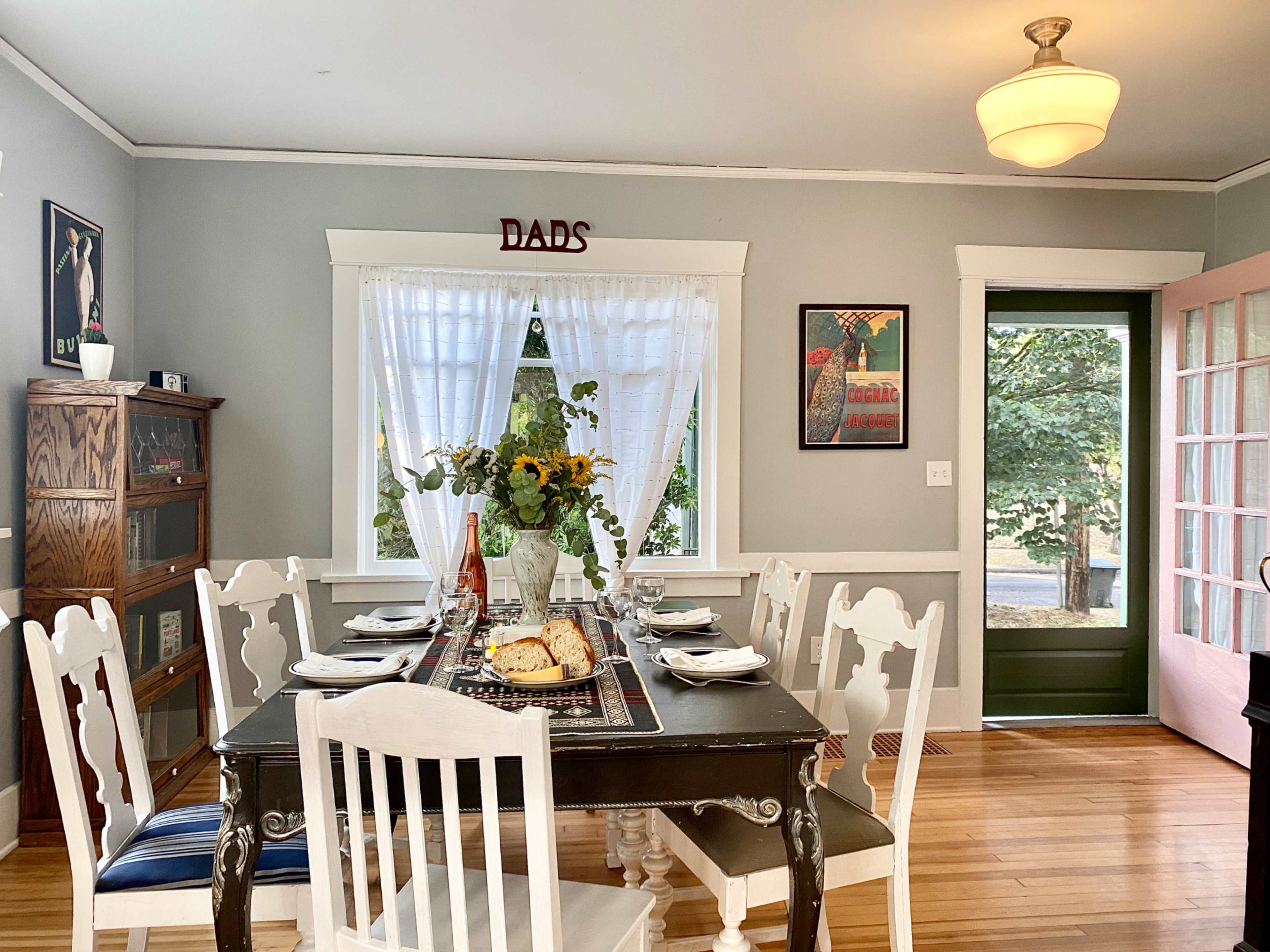 A dining room features a table set for a meal, surrounded by white chairs, with a vase of sunflowers on the table and a green door leading outside.