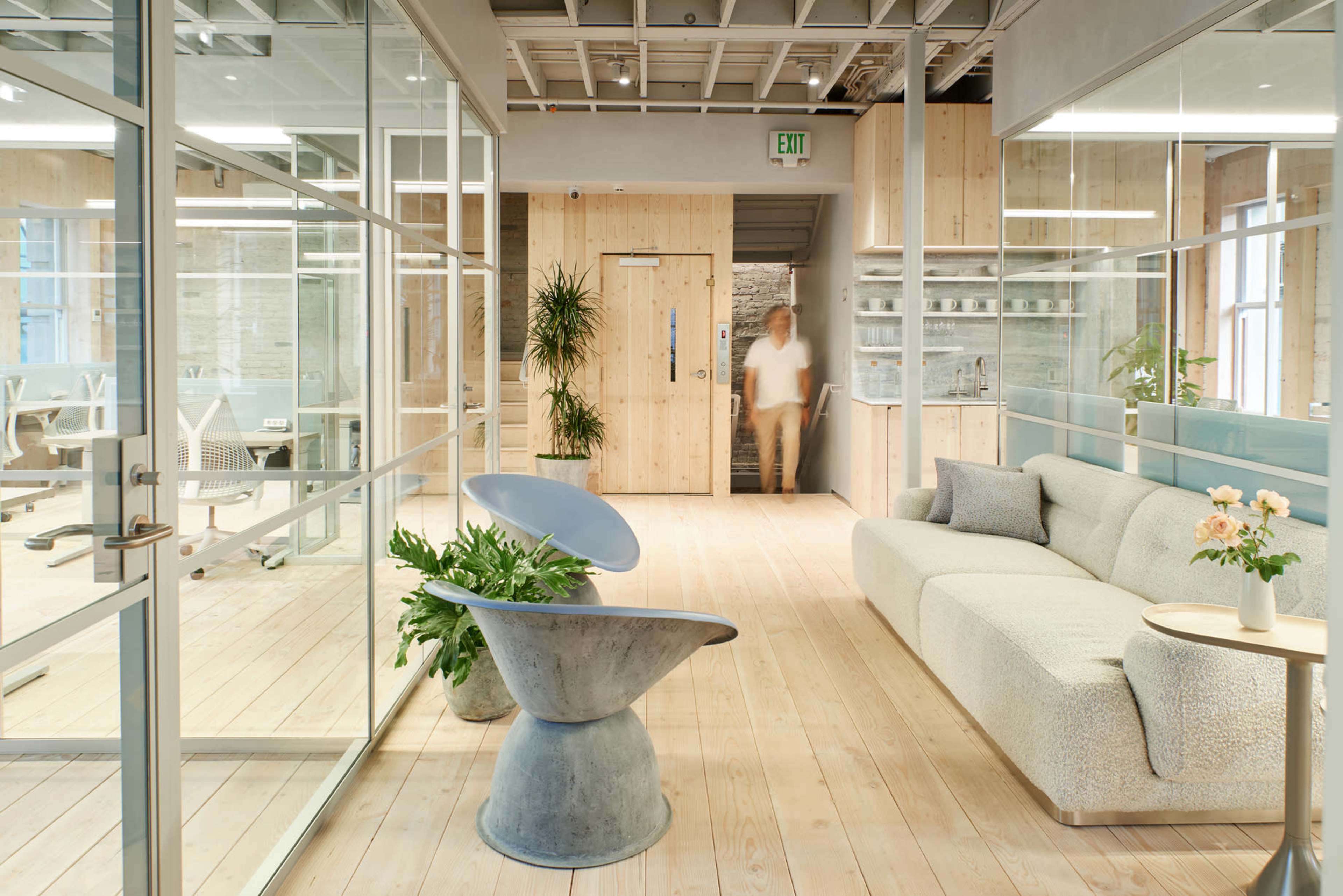 A modern office interior with glass walls, a light wooden floor, a minimalist sofa, and plants, leading to a doorway with a person walking through it.