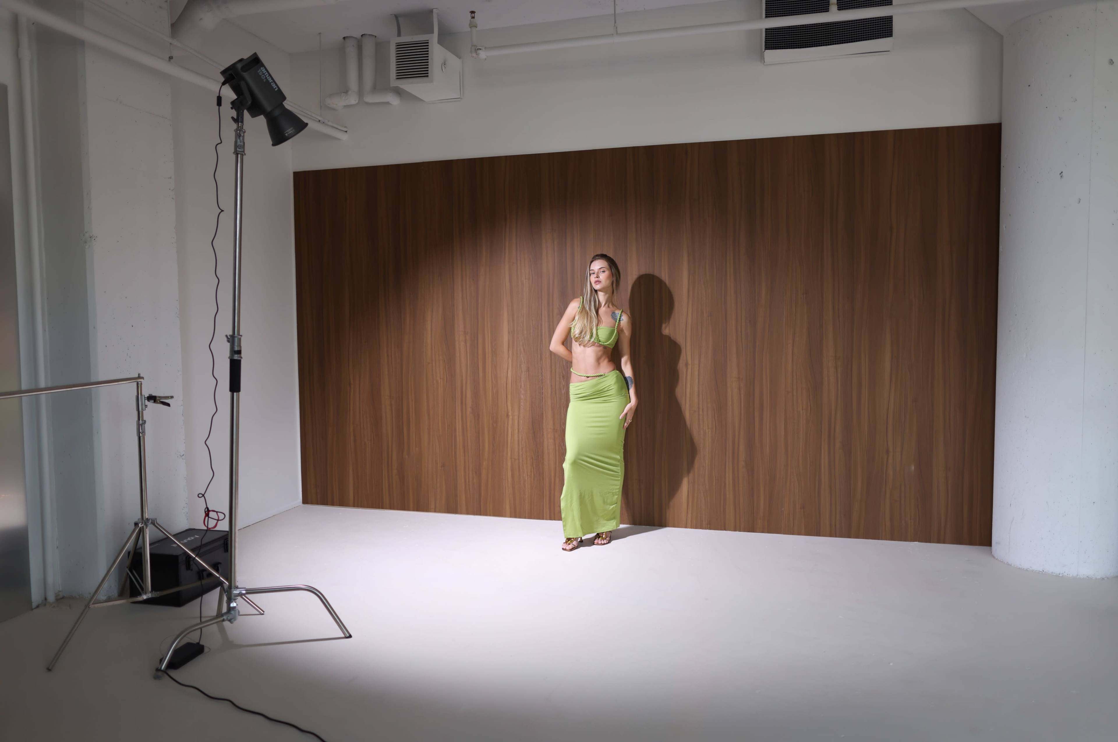 A model poses in a green outfit in front of a brown wooden backdrop under a spotlight in a studio.