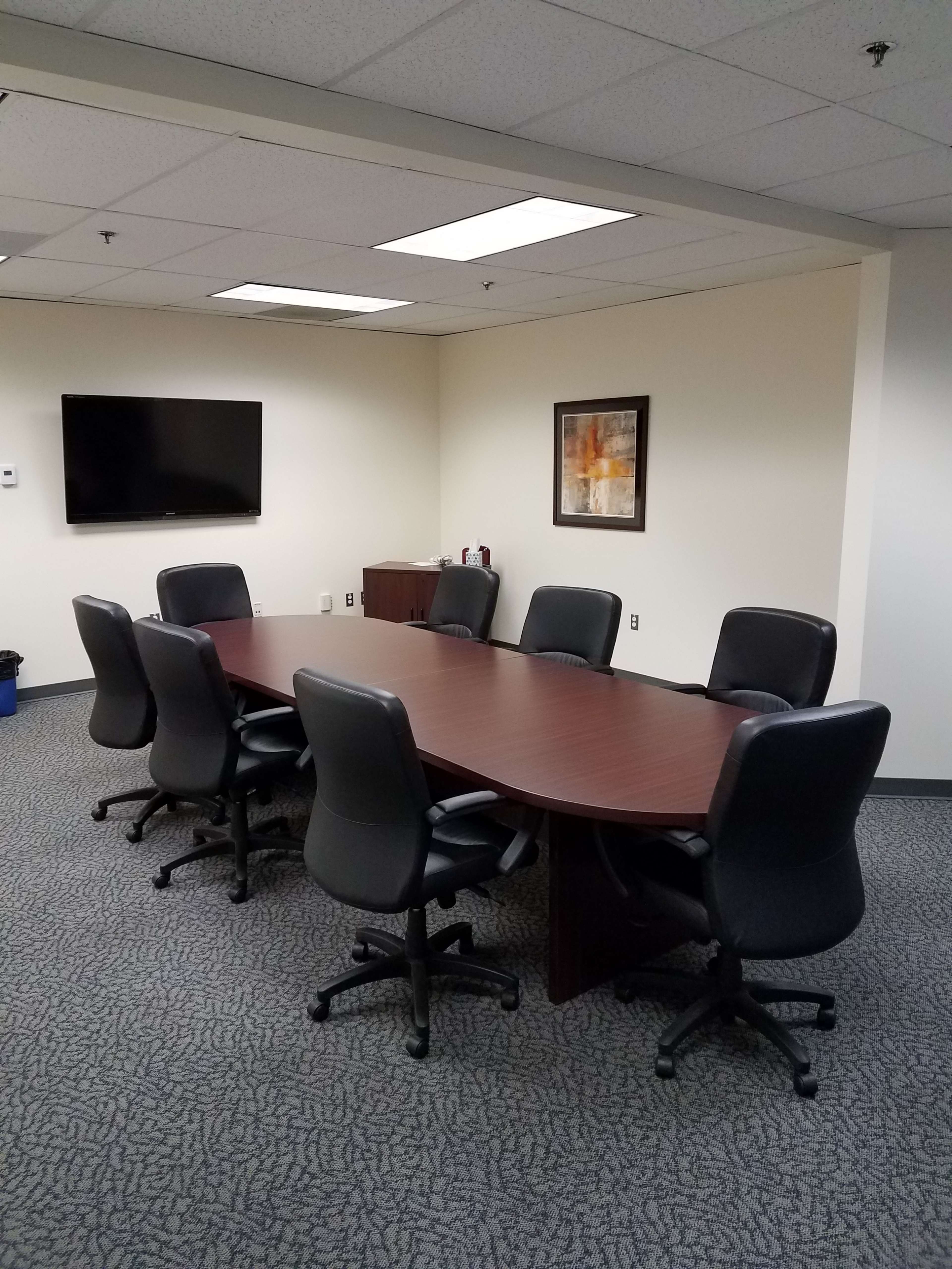 A conference room with a rectangular wooden table surrounded by eight black rolling chairs, a television mounted on the wall, and a piece of framed art.