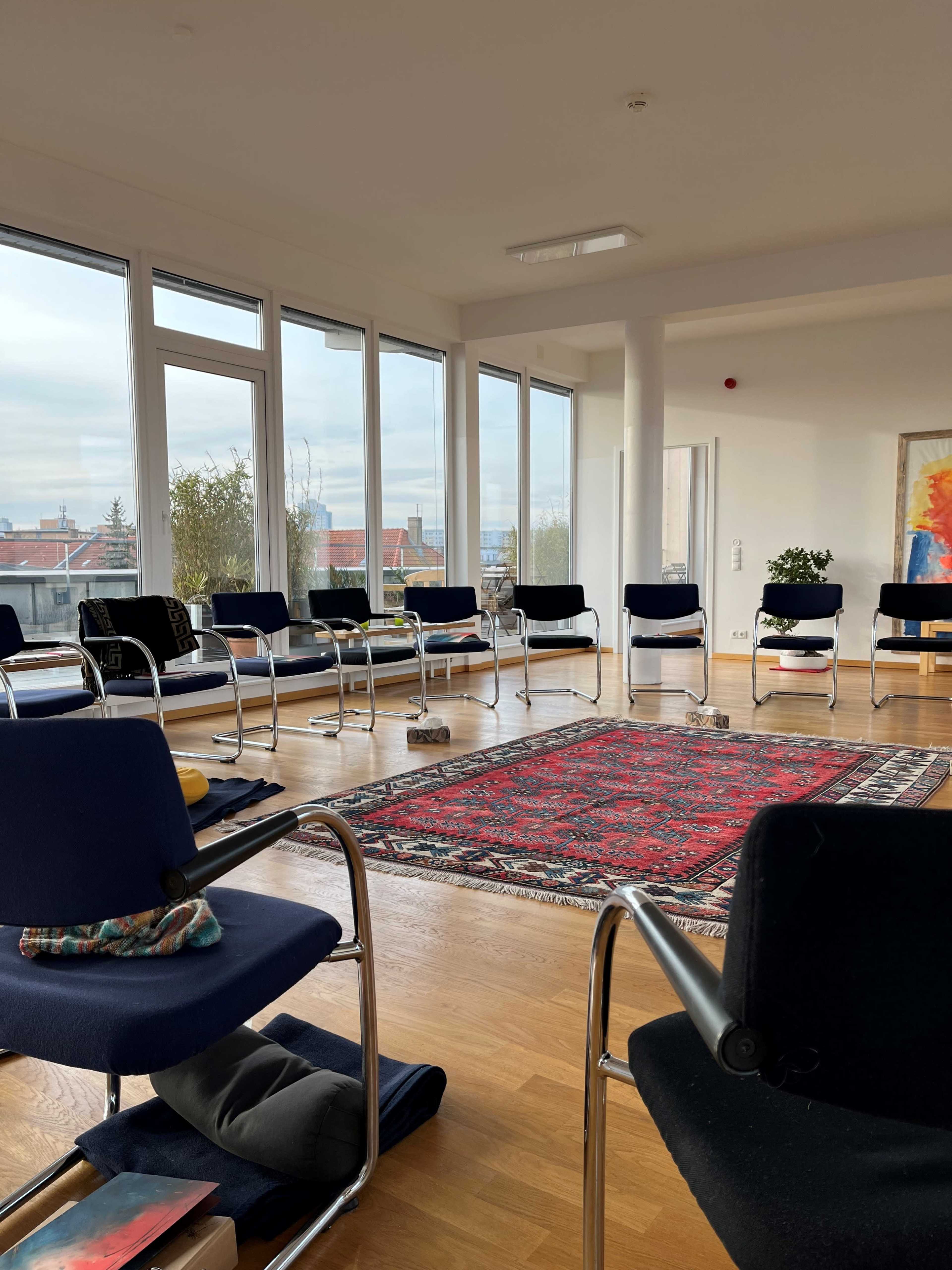 A circle of chairs surrounds a large, colorful rug in a well-lit, modern room with large windows.