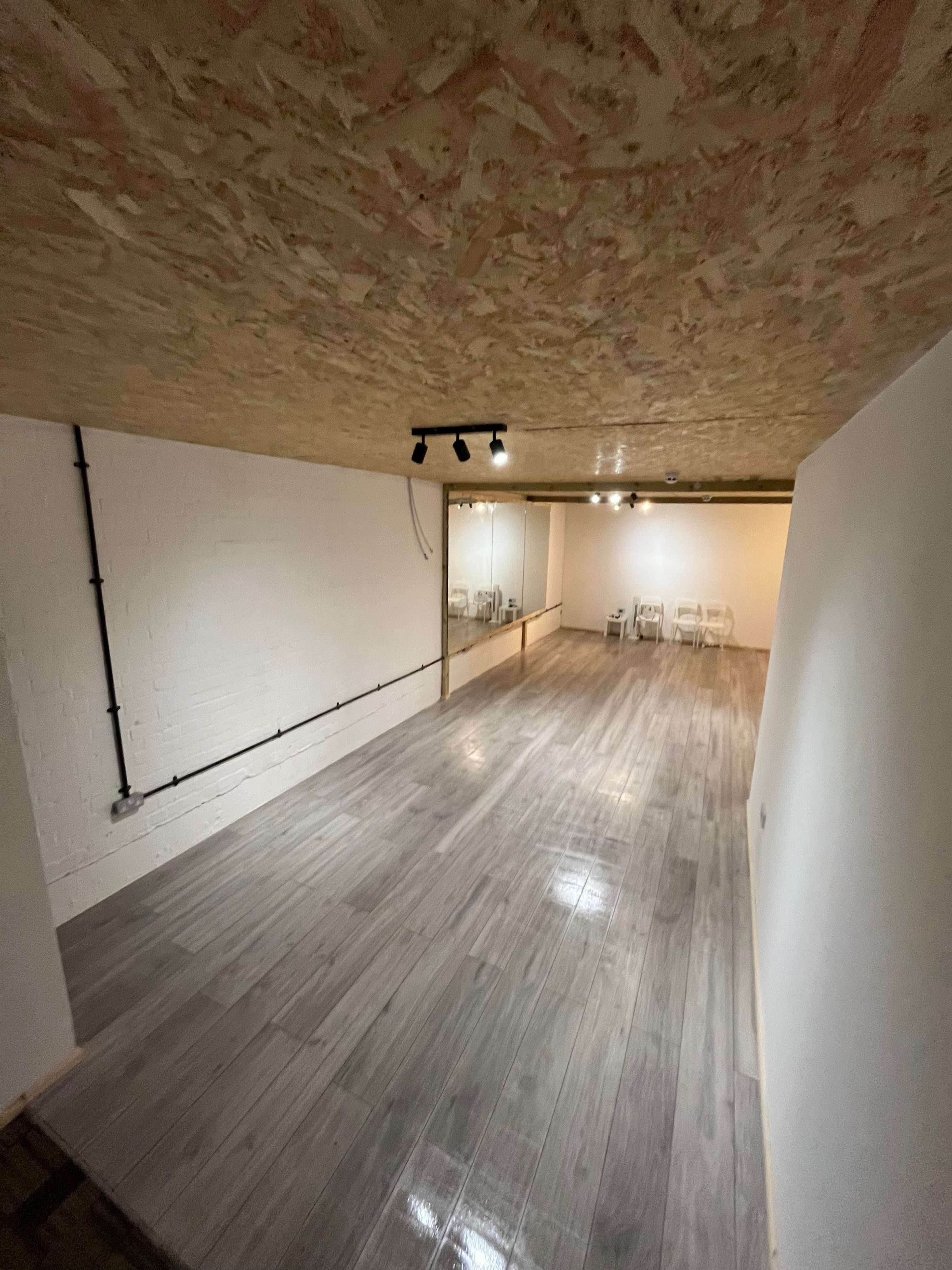 A spacious, empty room with a polished wooden floor, exposed ceiling, and a mirror on one wall.