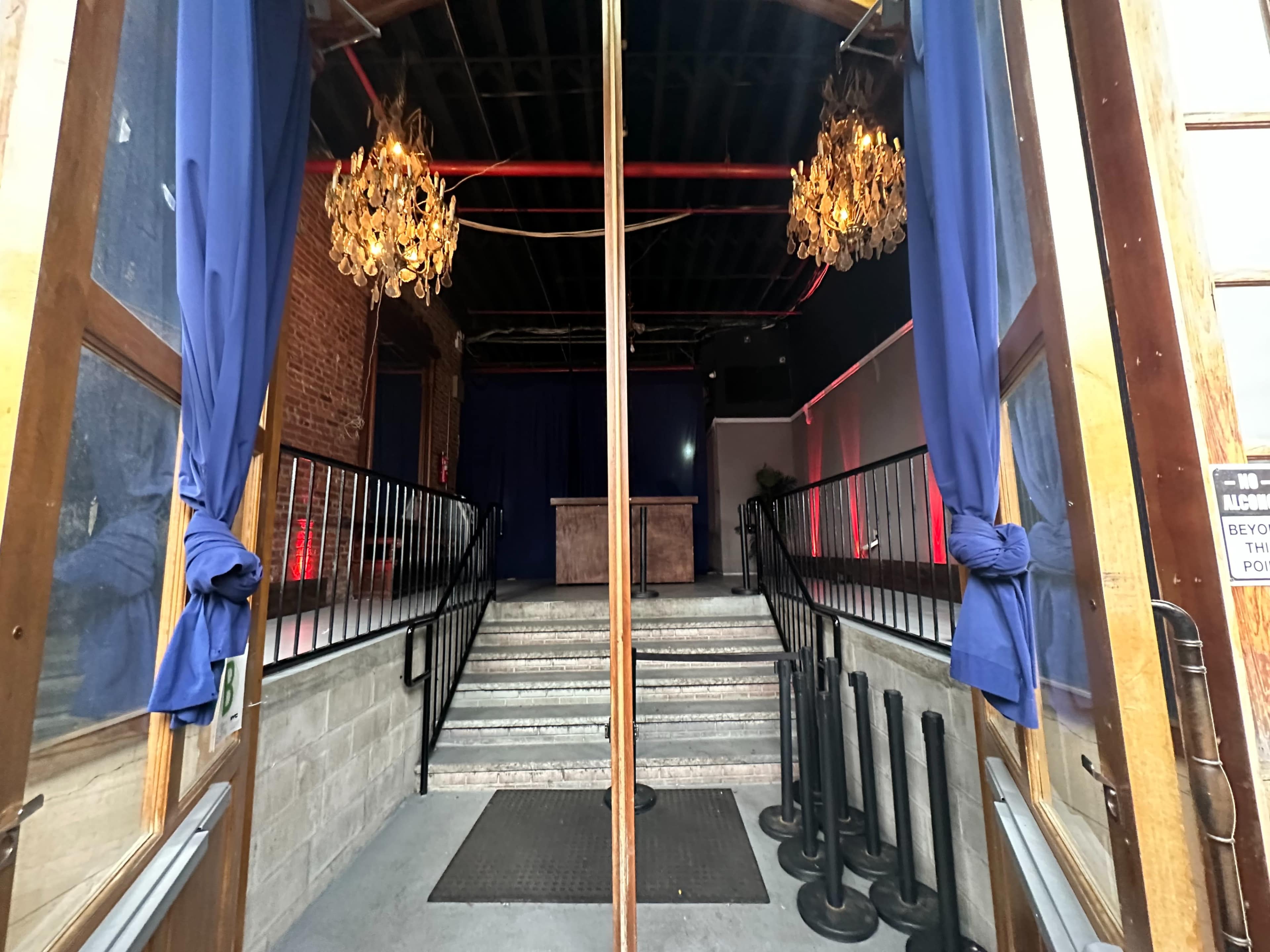 The entrance of a venue with wooden doors opened to reveal a staircase leading up to a stage area with two chandeliers and blue curtains.