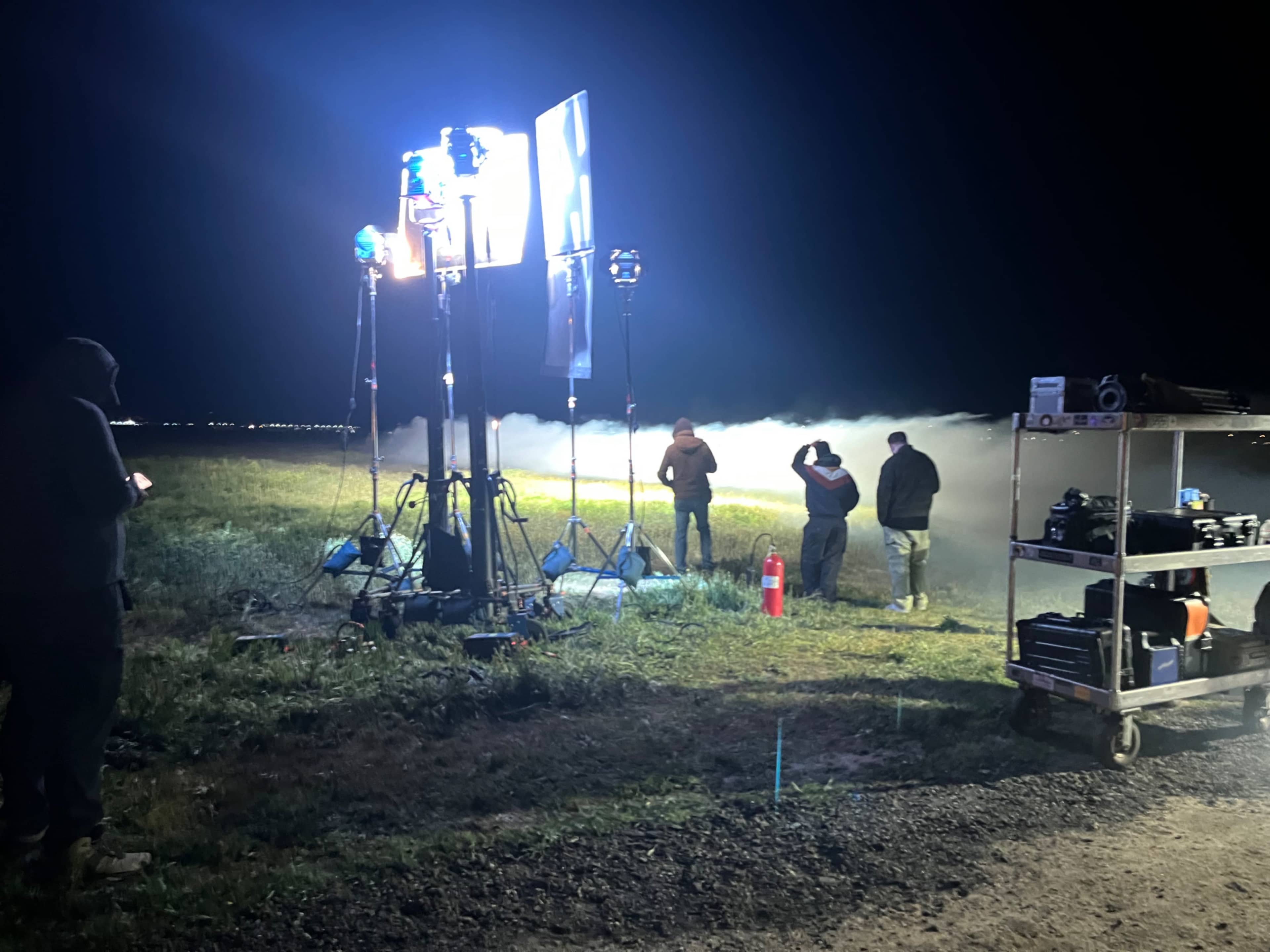 Crew members are set up on a dark field at night with lighting equipment illuminating the area, while smoke effects are being used in the background.