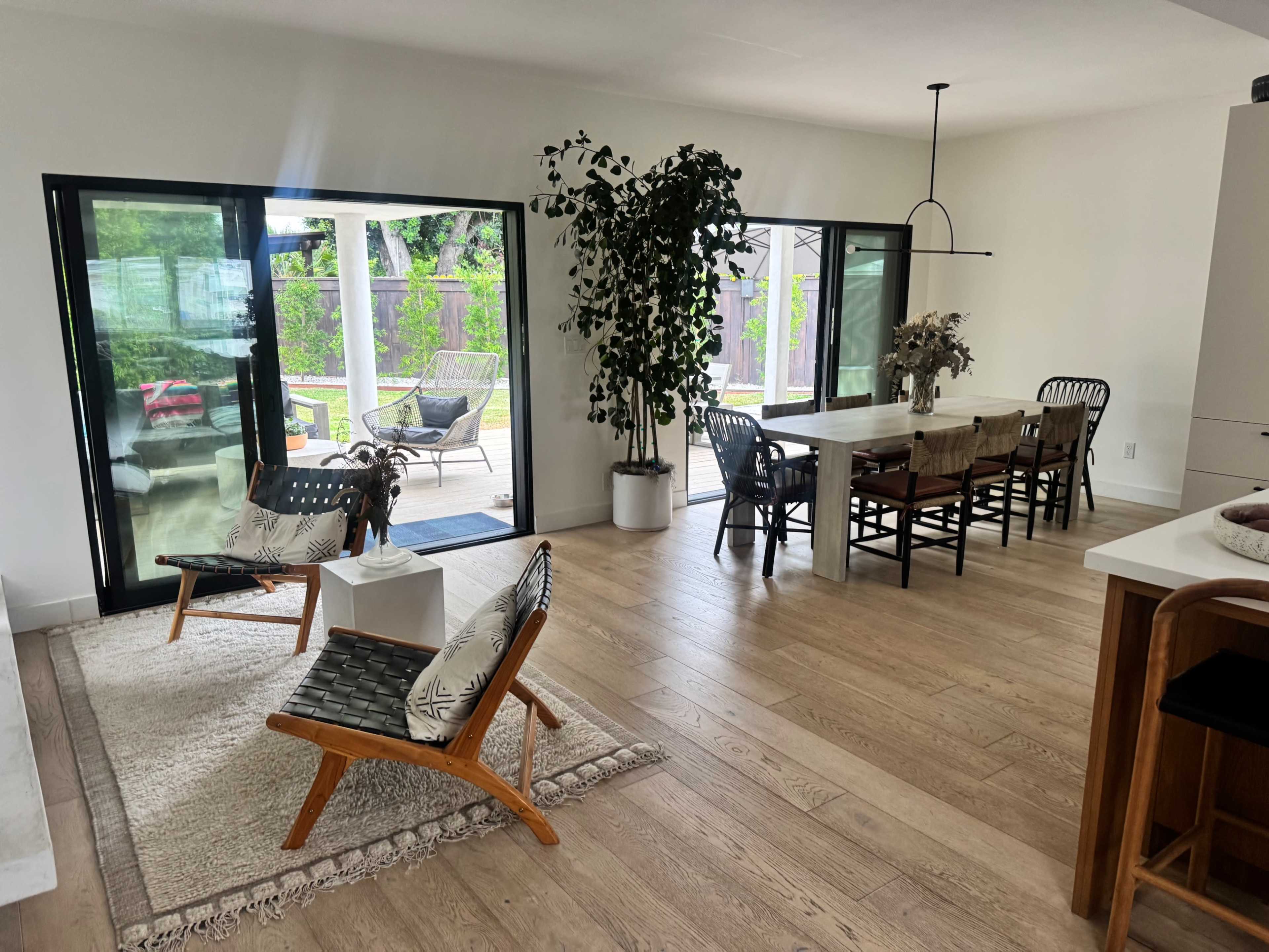 The image shows a modern open-concept living and dining area featuring wooden flooring, a large dining table, and lounge chairs near sliding glass doors that lead to an outdoor patio.