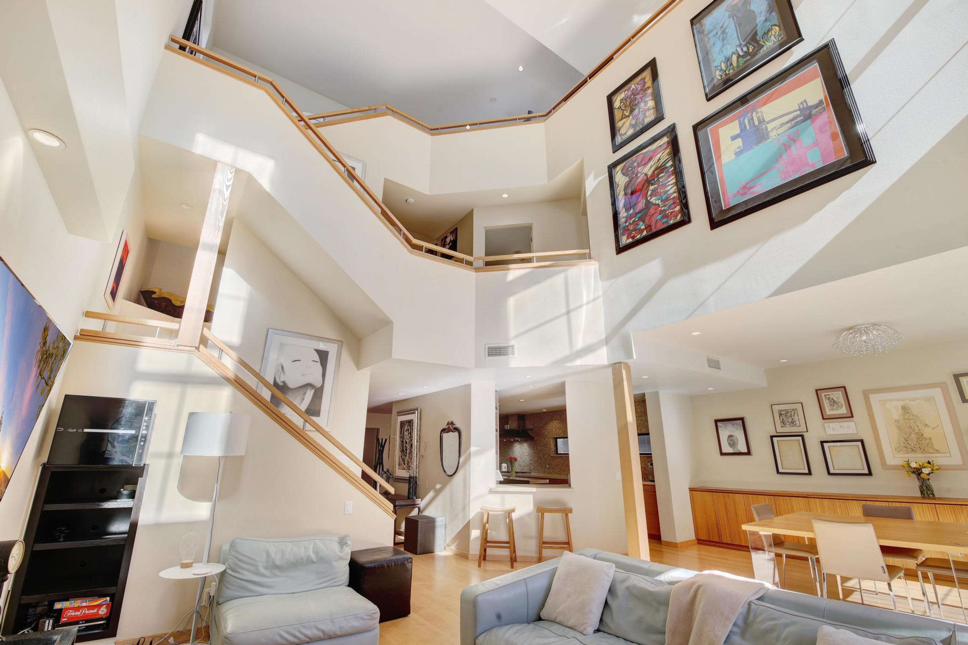 A modern, multi-level living space with large windows, wooden stairs, and various framed artworks on the walls.