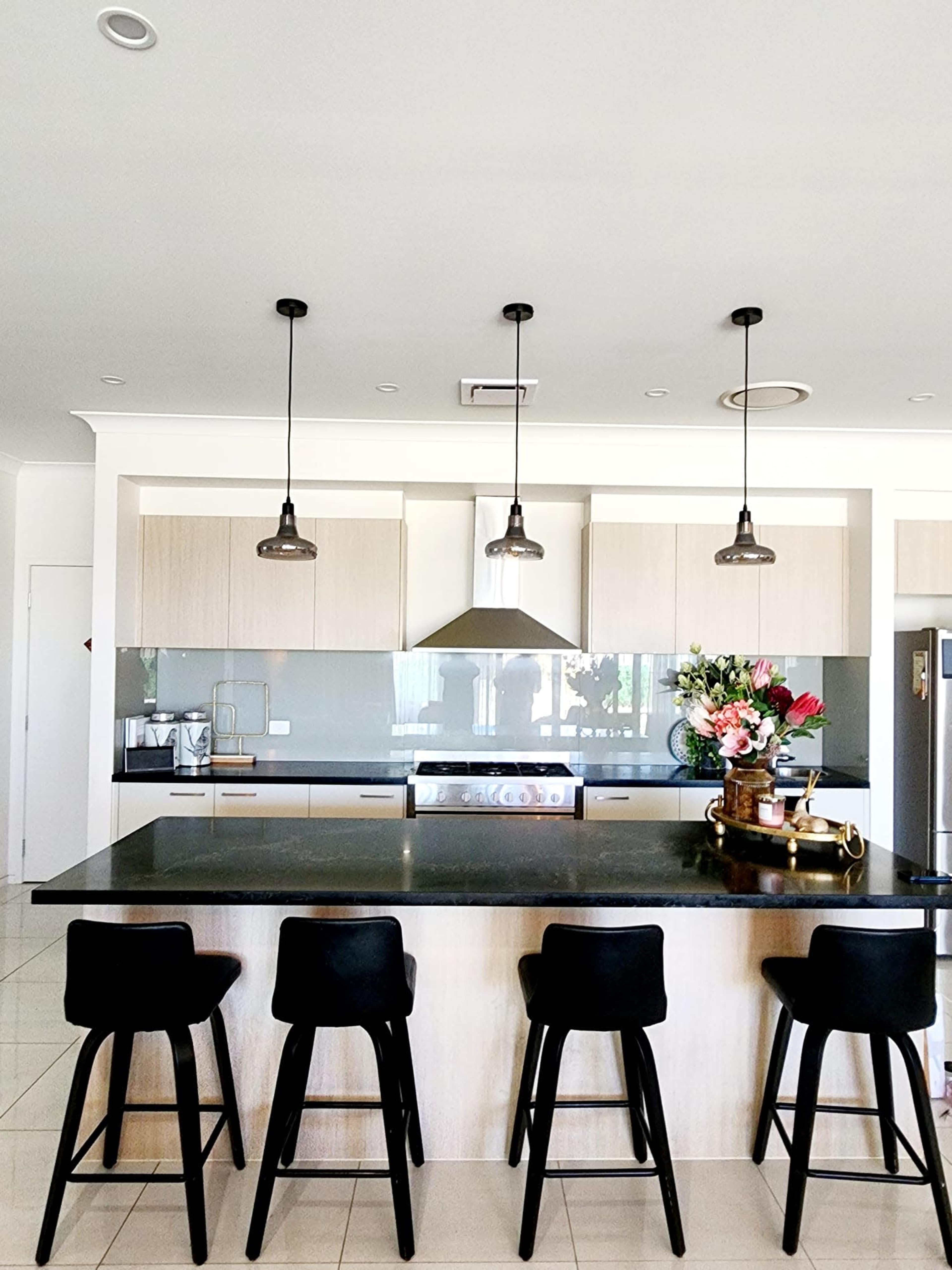 A modern kitchen features a dark countertop and light wooden cabinets, with four black bar stools positioned at a large island and pendant lights hanging above.