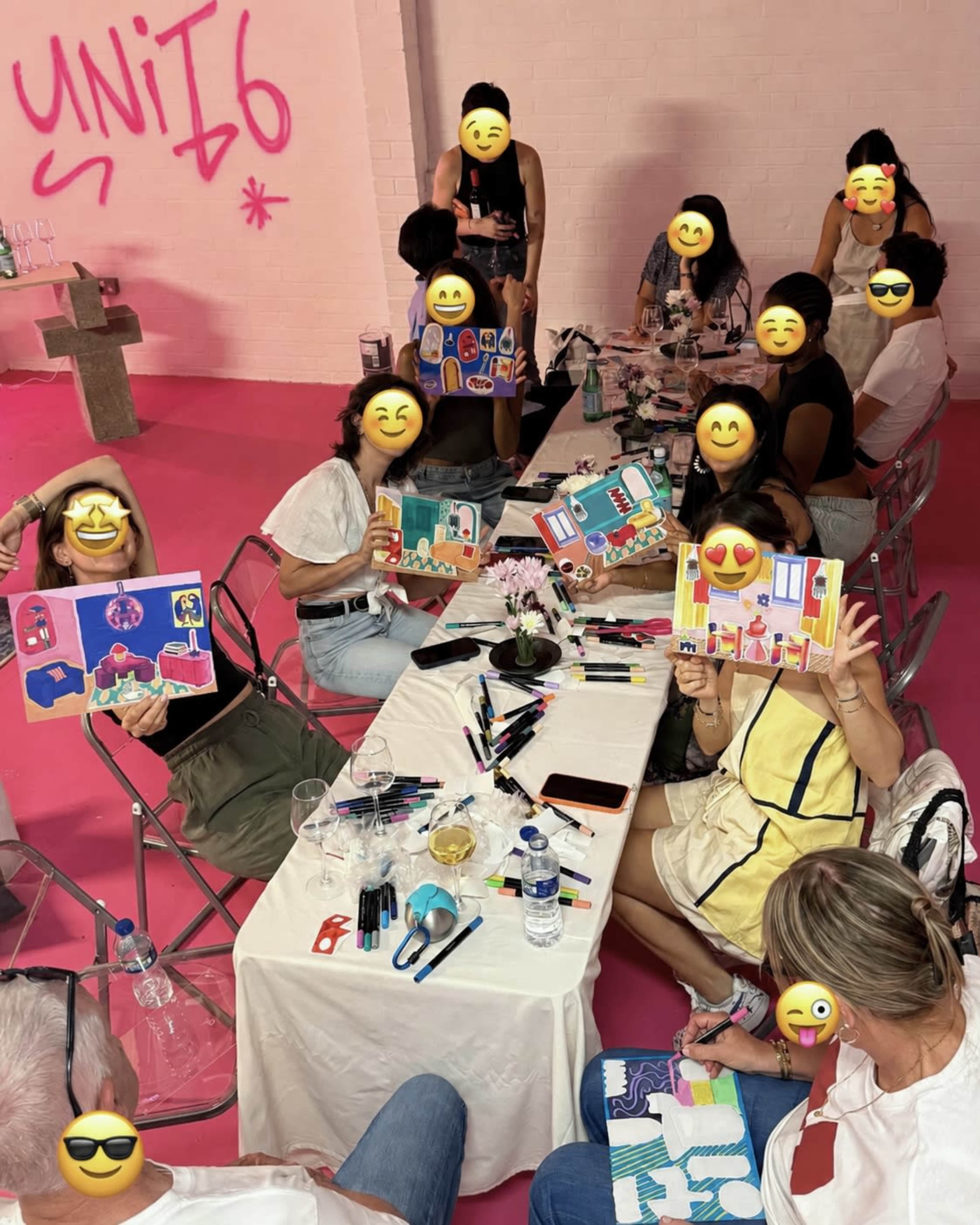 A group of people is sitting around a table engaged in an art activity, displaying their colorful artwork while surrounded by pink decor.