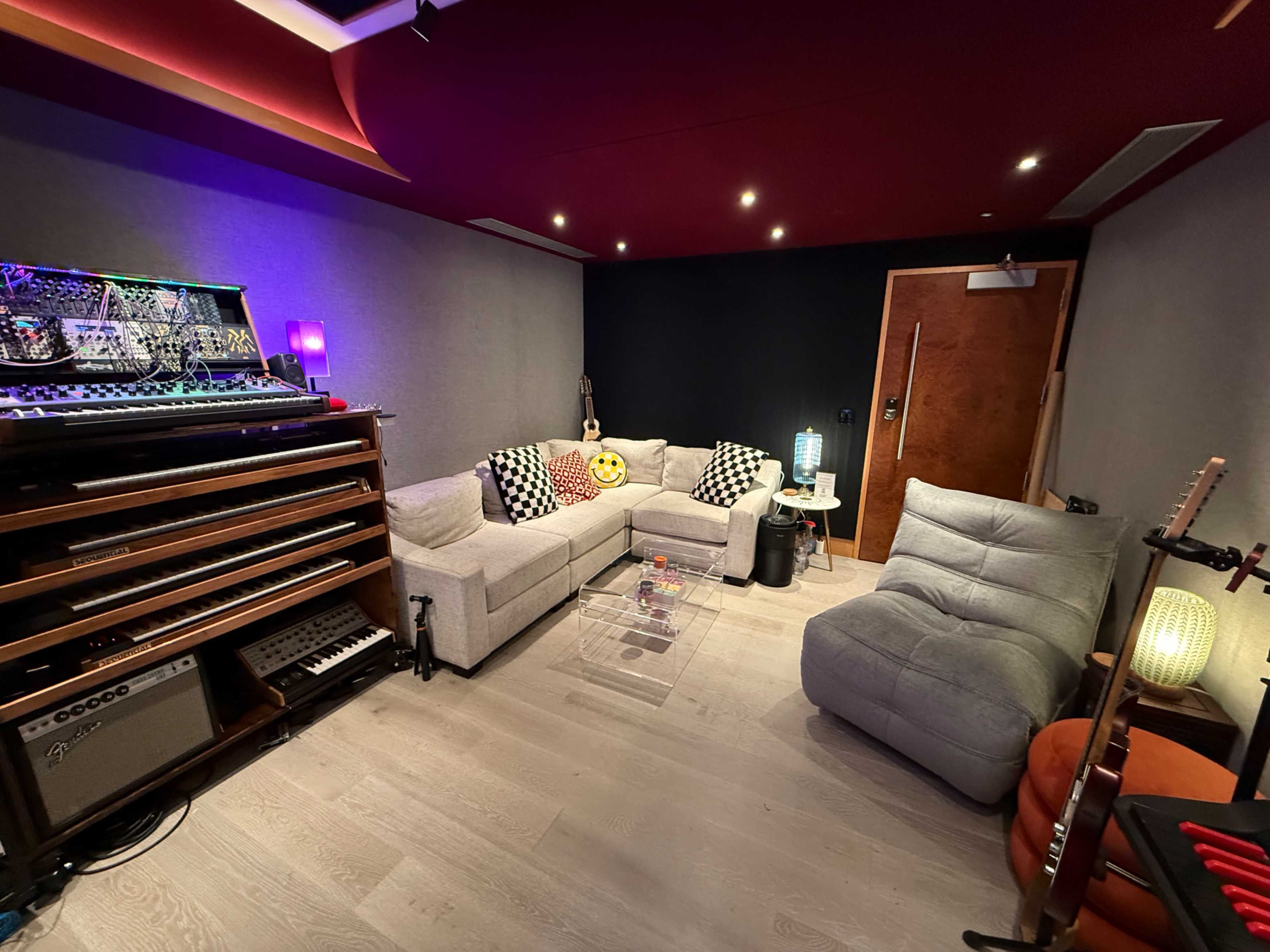 The image shows a cozy room equipped with a sound mixing setup, featuring a sectional sofa, various musical instruments, and decorative lighting.