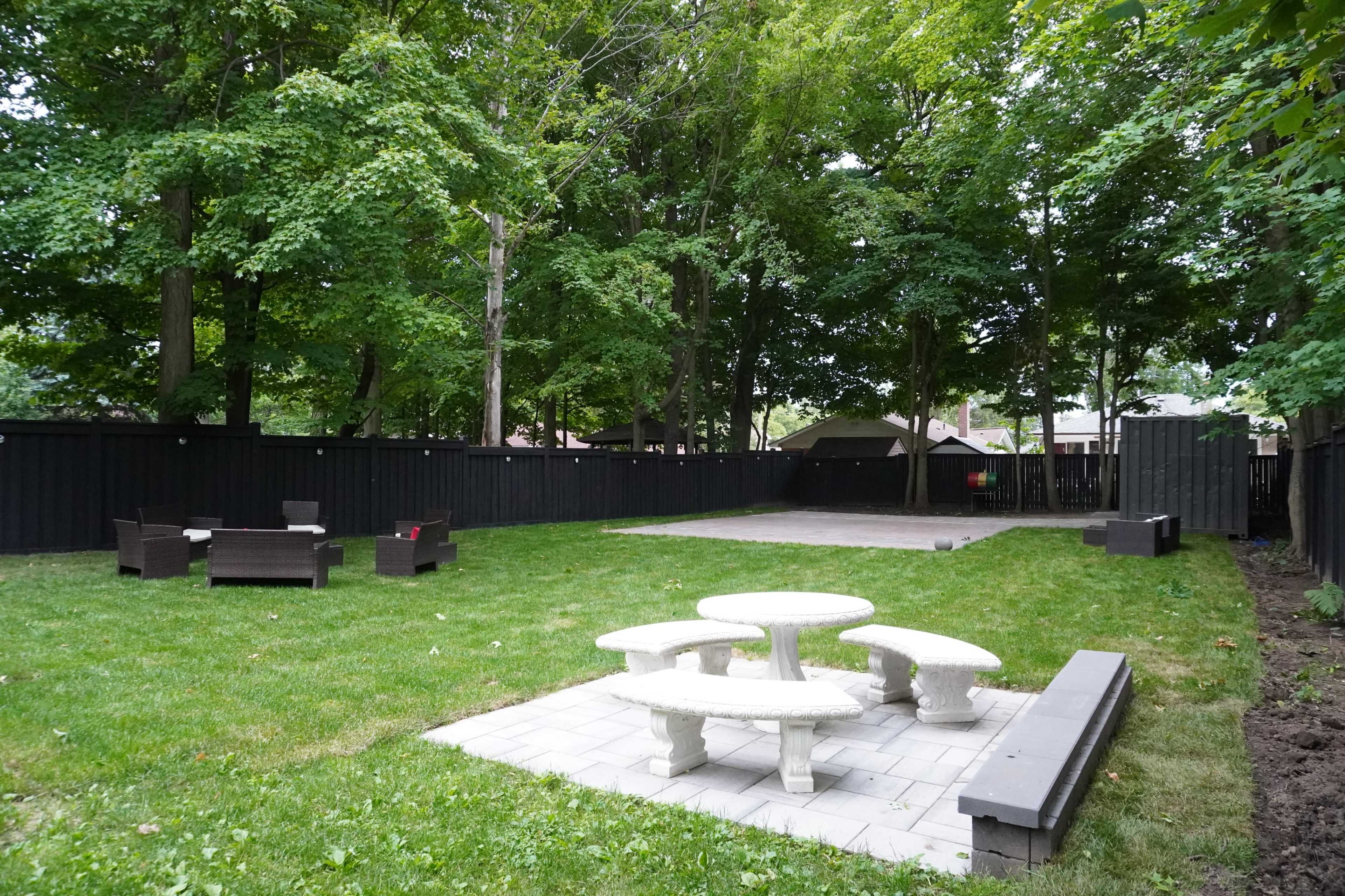 The image shows a spacious backyard with a stone table and benches, a seating area made of dark wicker furniture, and tall trees surrounding a grassy lawn.