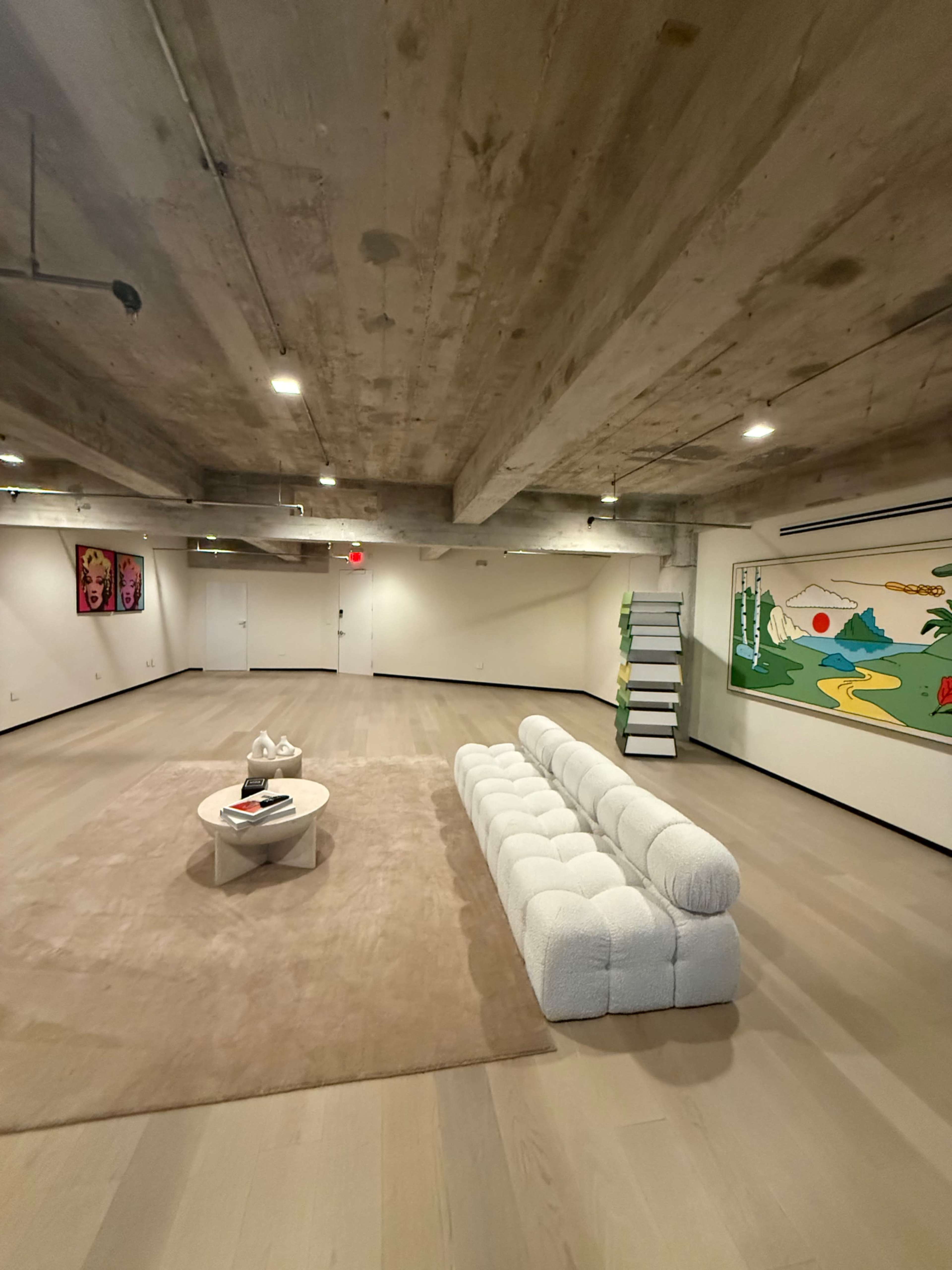 The image shows a spacious, minimalist interior with a light-colored floor, a large area rug, a white curved sofa, a round coffee table, and colorful artwork on the walls.