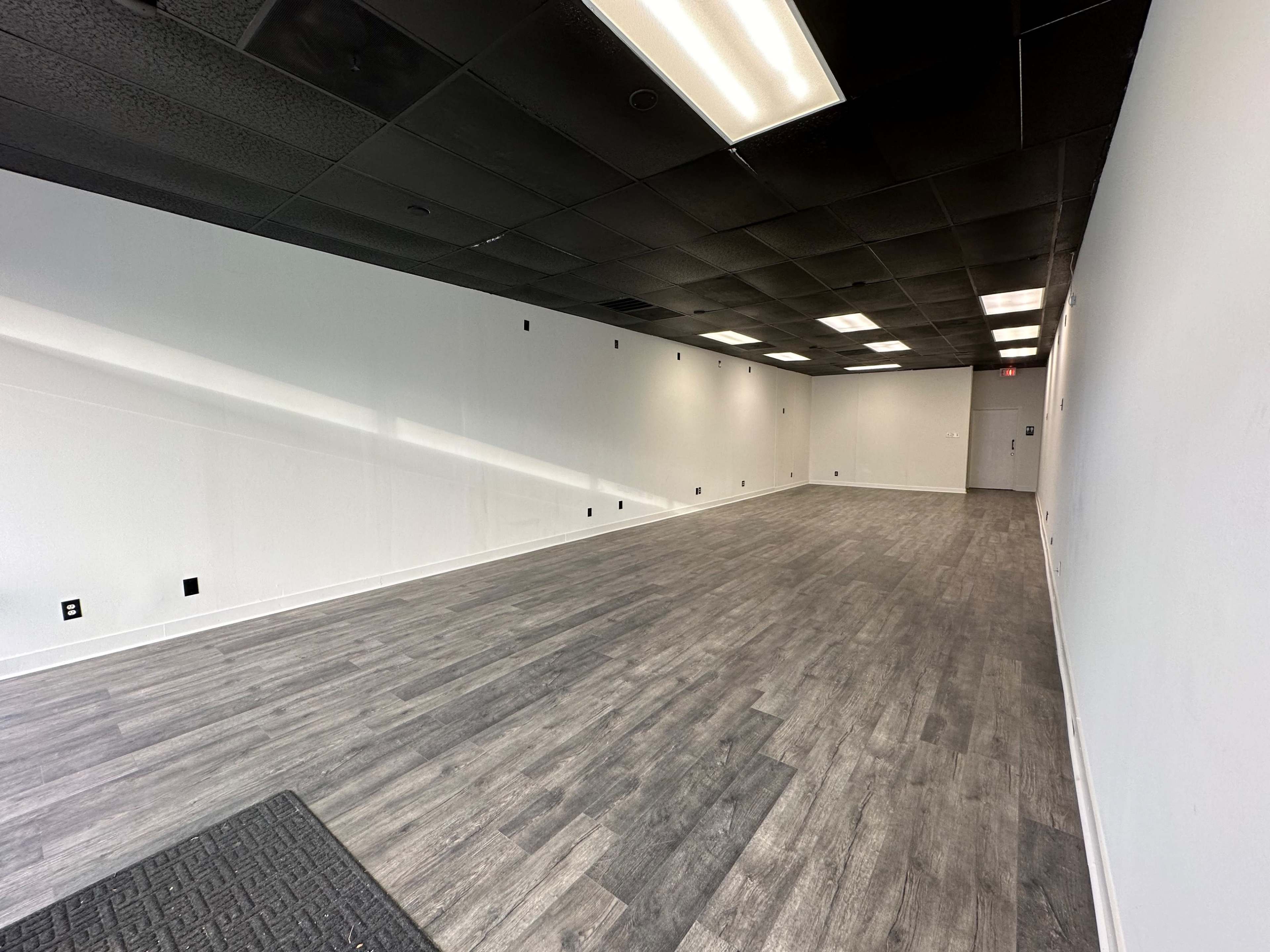 A spacious, empty room with gray wooden flooring and white walls, illuminated by overhead lights.