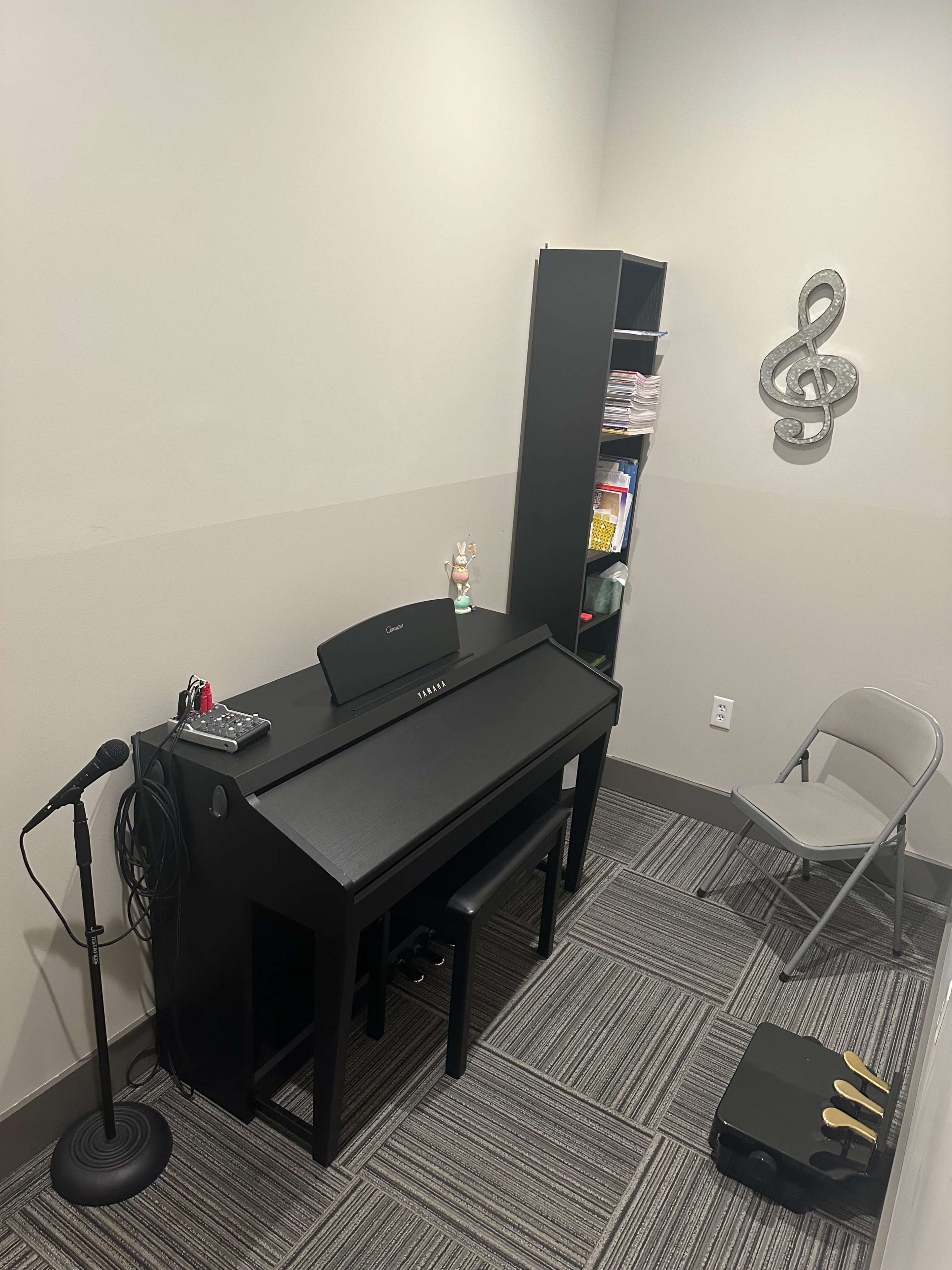 The image shows a small music practice room featuring a black digital piano, a microphone stand, a gray folding chair, and a bookshelf filled with music materials.
