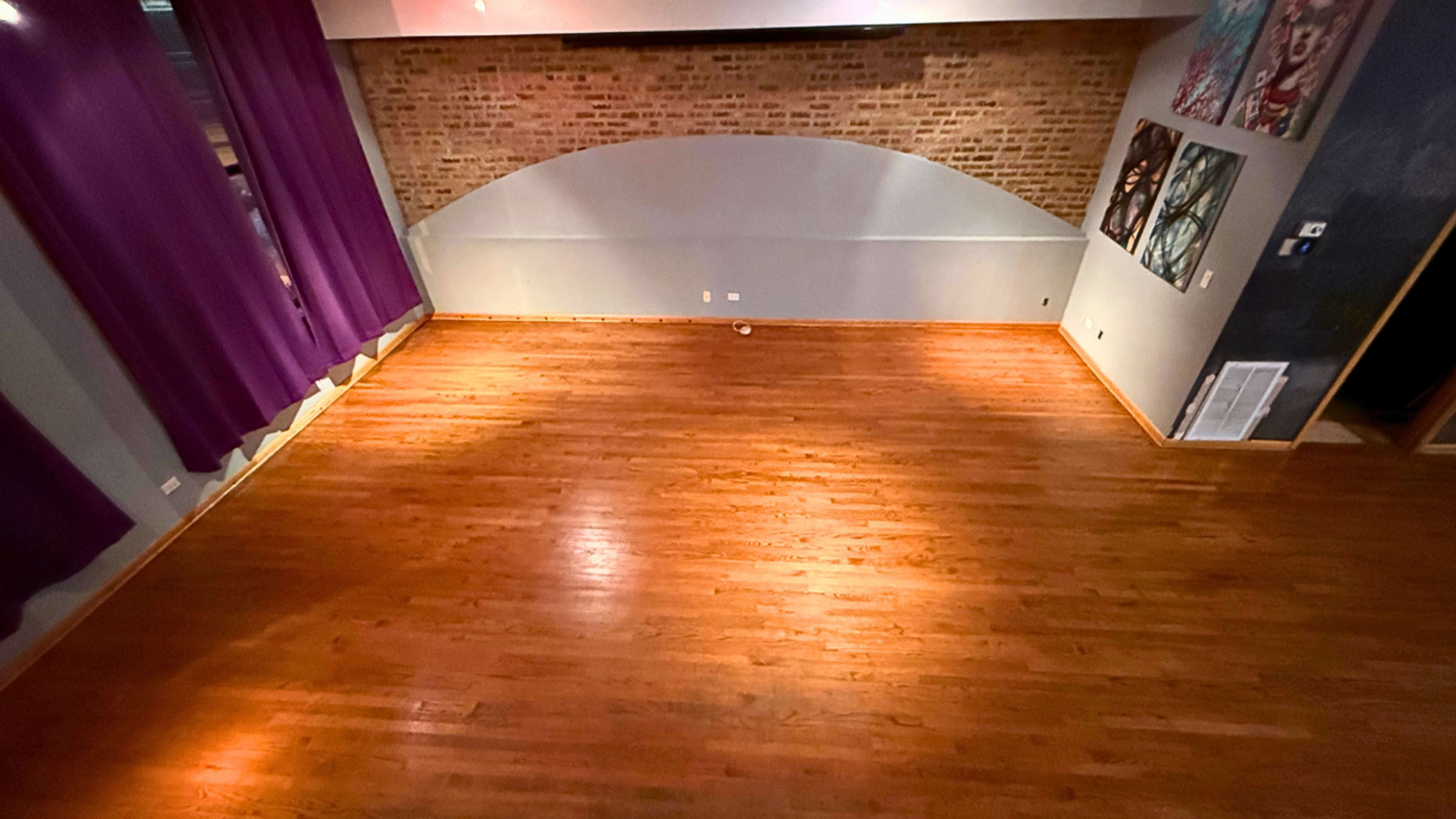 The image shows a spacious, empty room with polished wooden floors and brick walls, illuminated by soft overhead lighting.