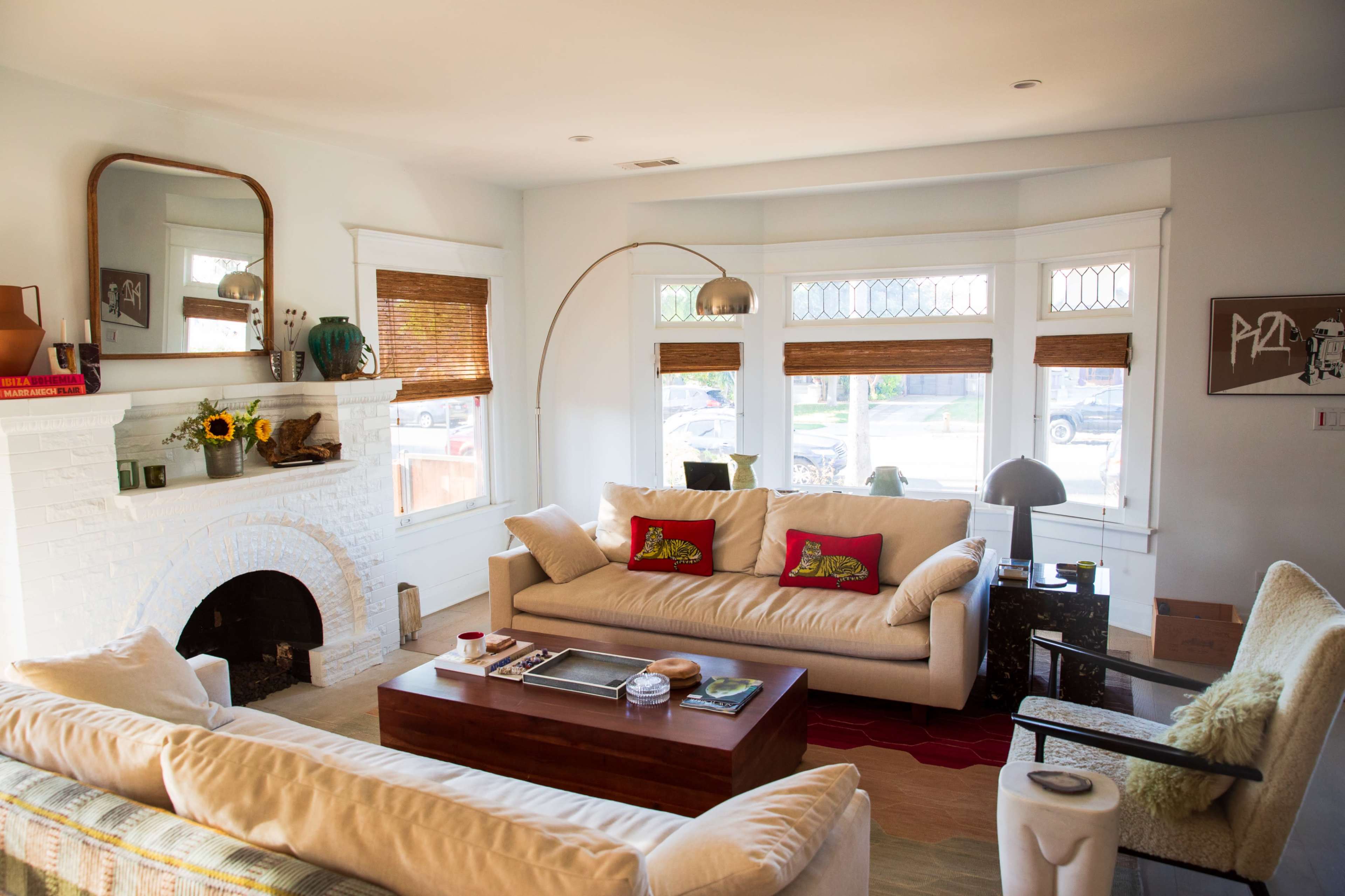 The living room features two beige sofas, a wooden coffee table, and large windows with bamboo blinds, all centered around a white brick fireplace.
