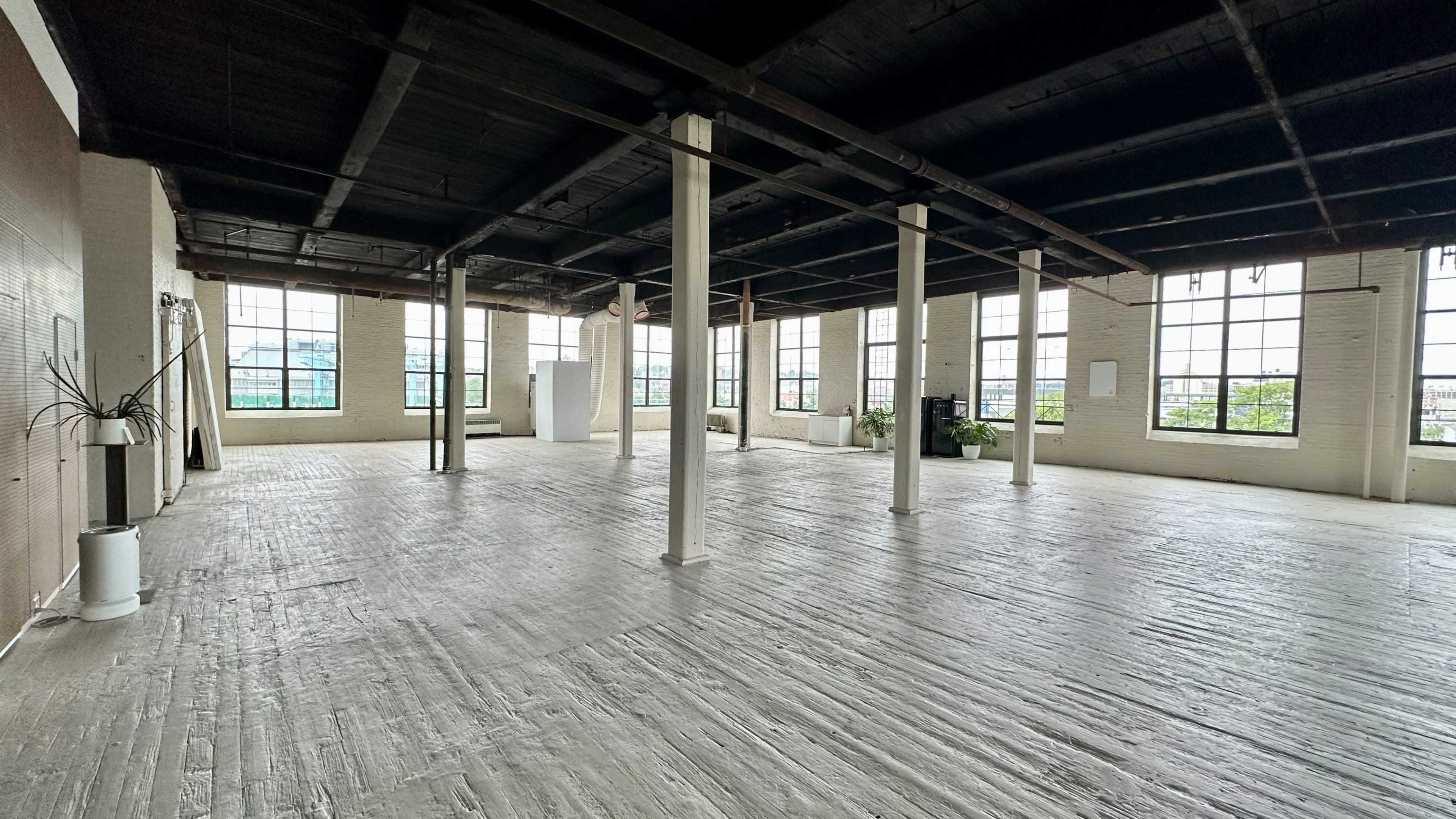 A wide, empty industrial-style room with large windows and wooden flooring.