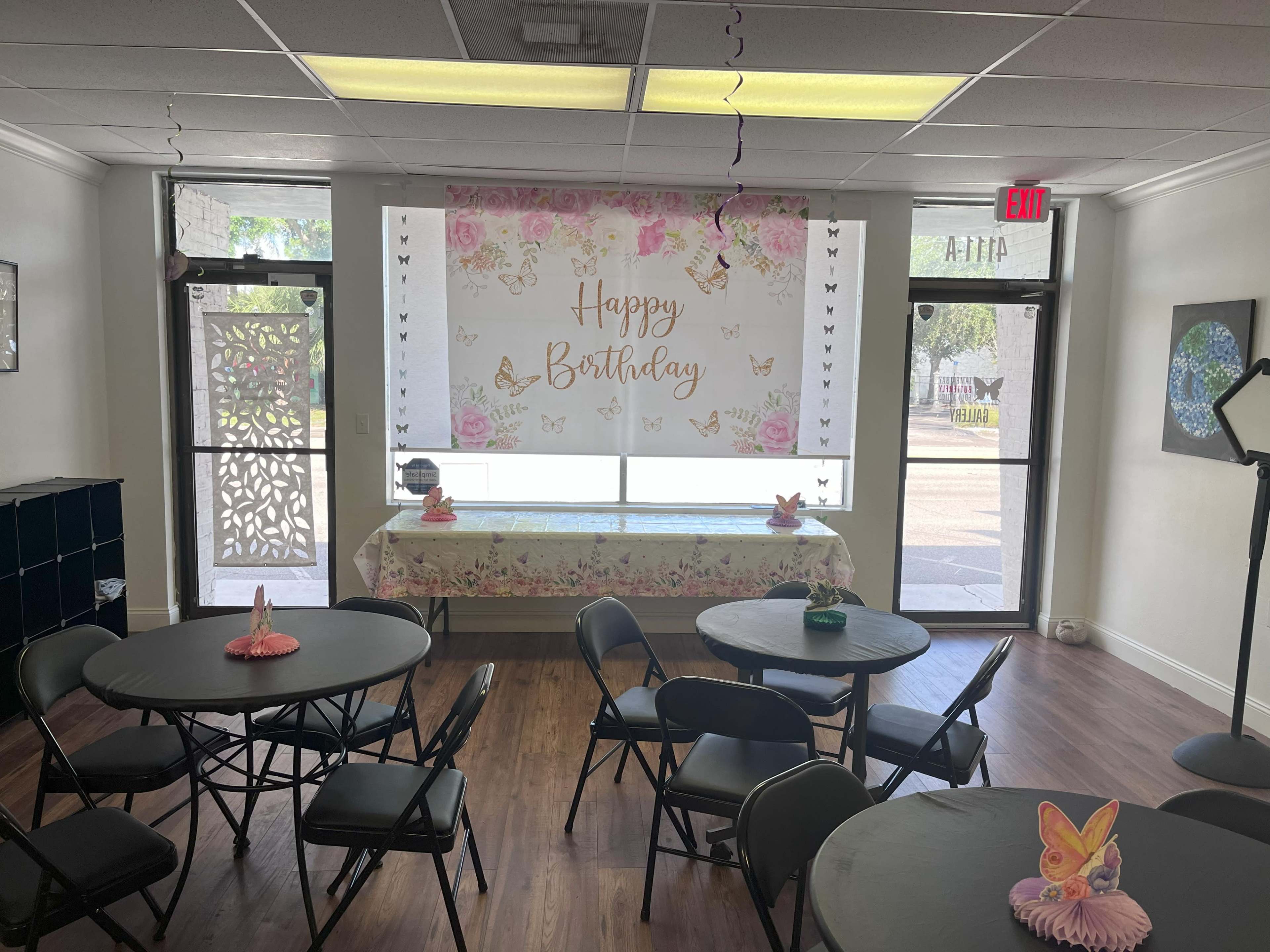 The image shows a decorated room with black round tables and a banner that reads "Happy Birthday" on the wall, along with a table covered in floral fabric and party decorations.