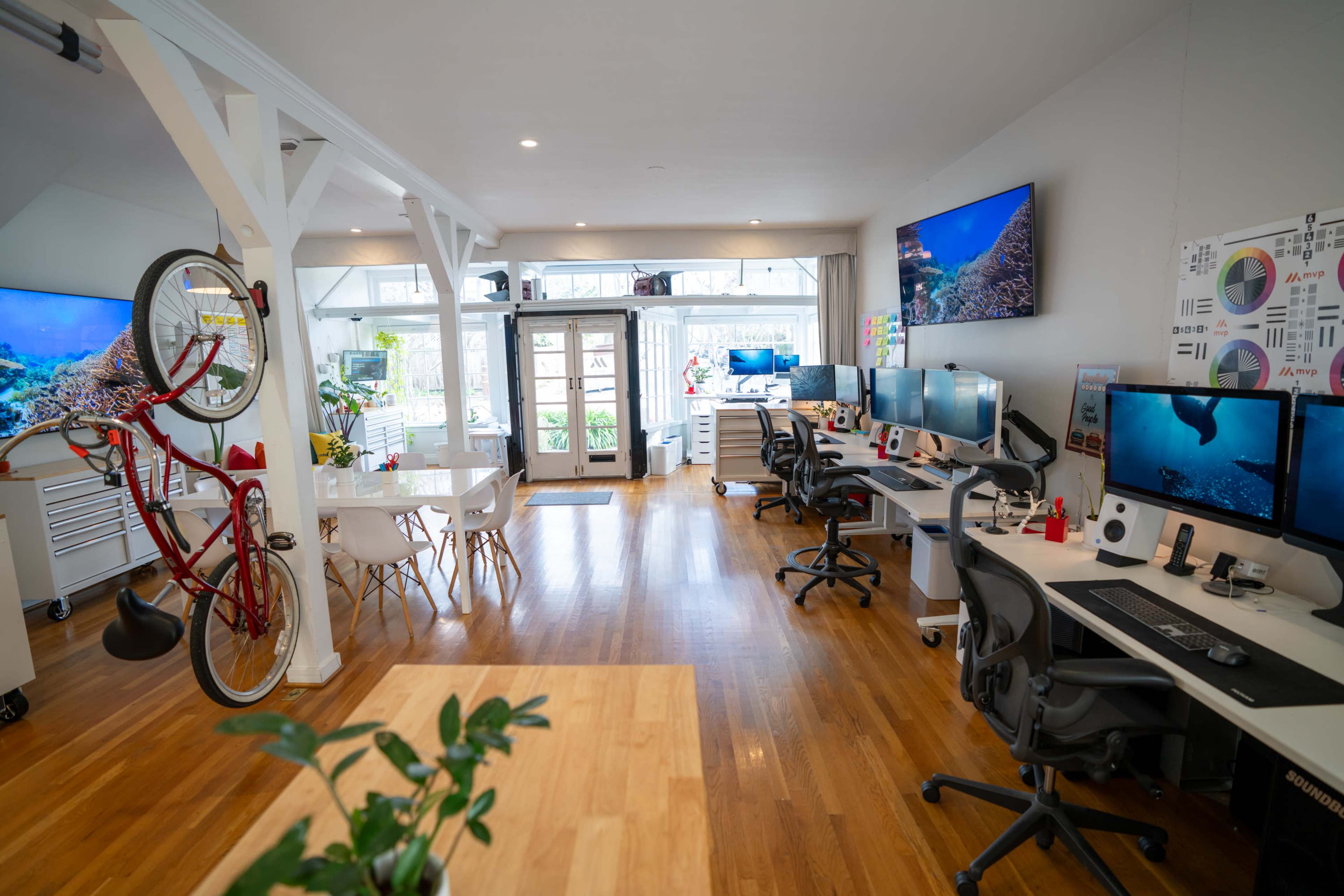 The image shows a modern office space featuring multiple workstations with computer monitors, a dining area with a table, and a bicycle hanging on the wall.