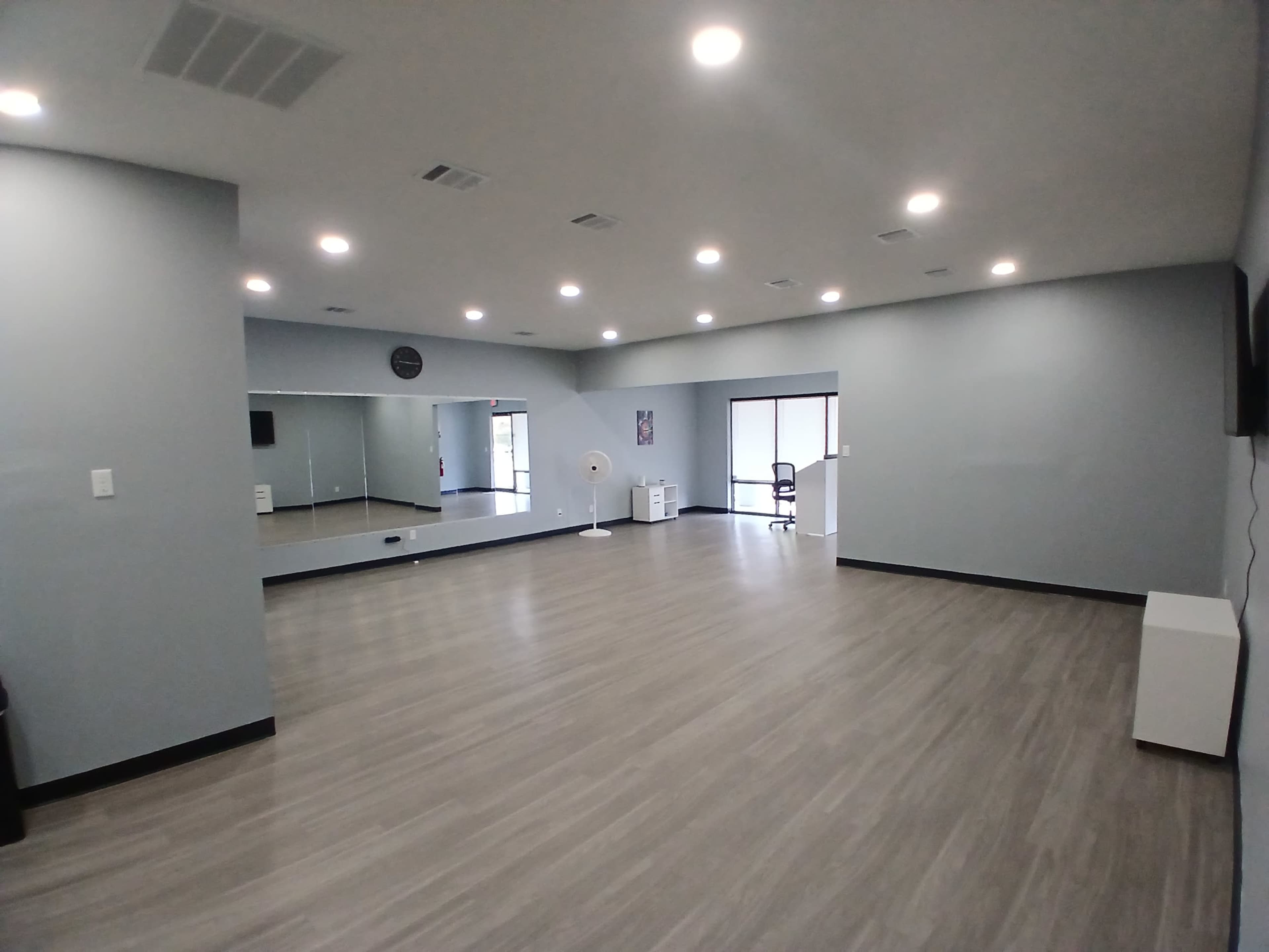 The image shows a spacious, well-lit room with a smooth wooden floor, featuring a large wall mirror and a window that allows natural light to enter.