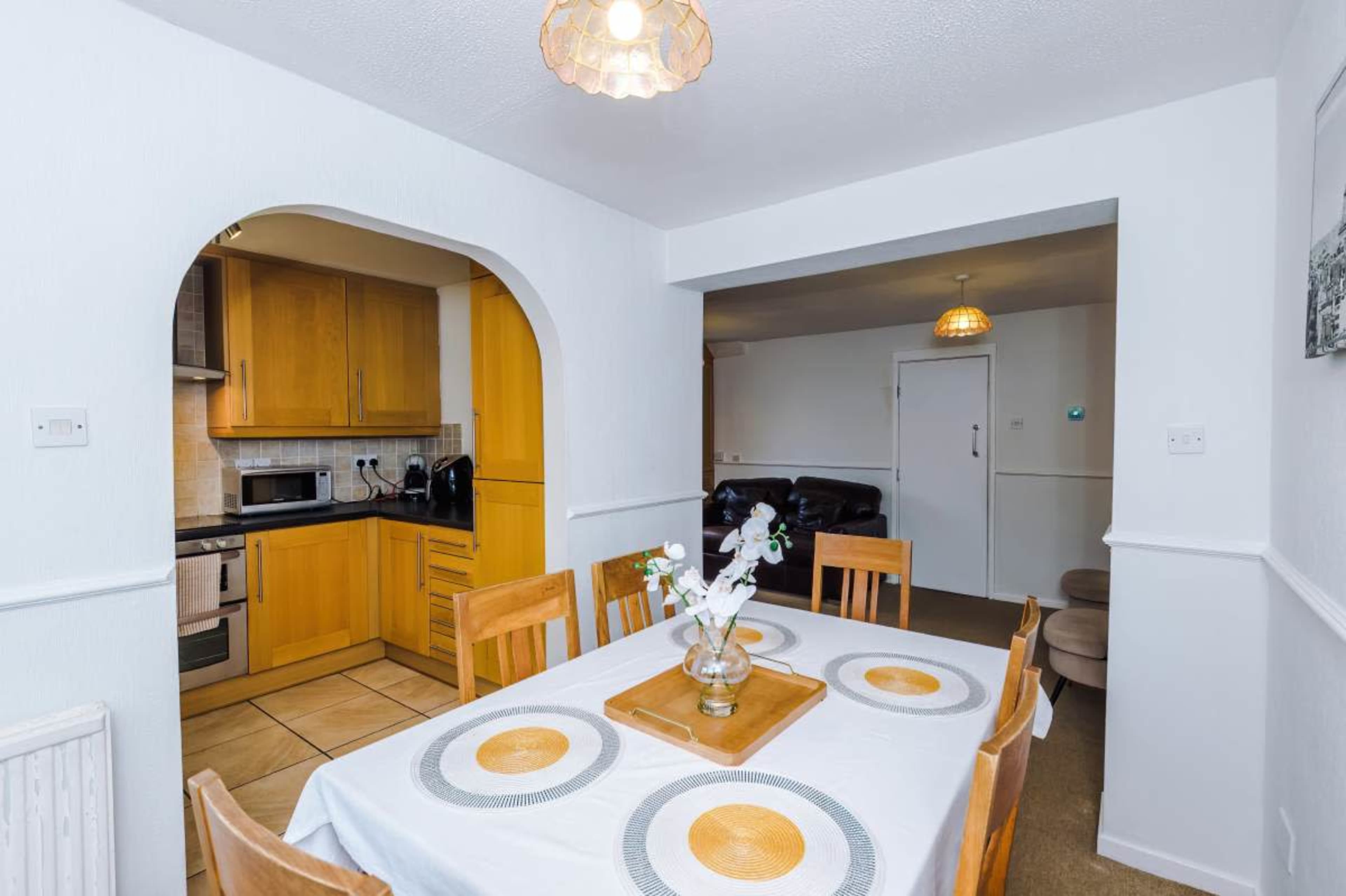The image shows a dining area with a table featuring a white tablecloth and a vase of flowers, adjacent to a kitchen with wooden cabinets and modern appliances.